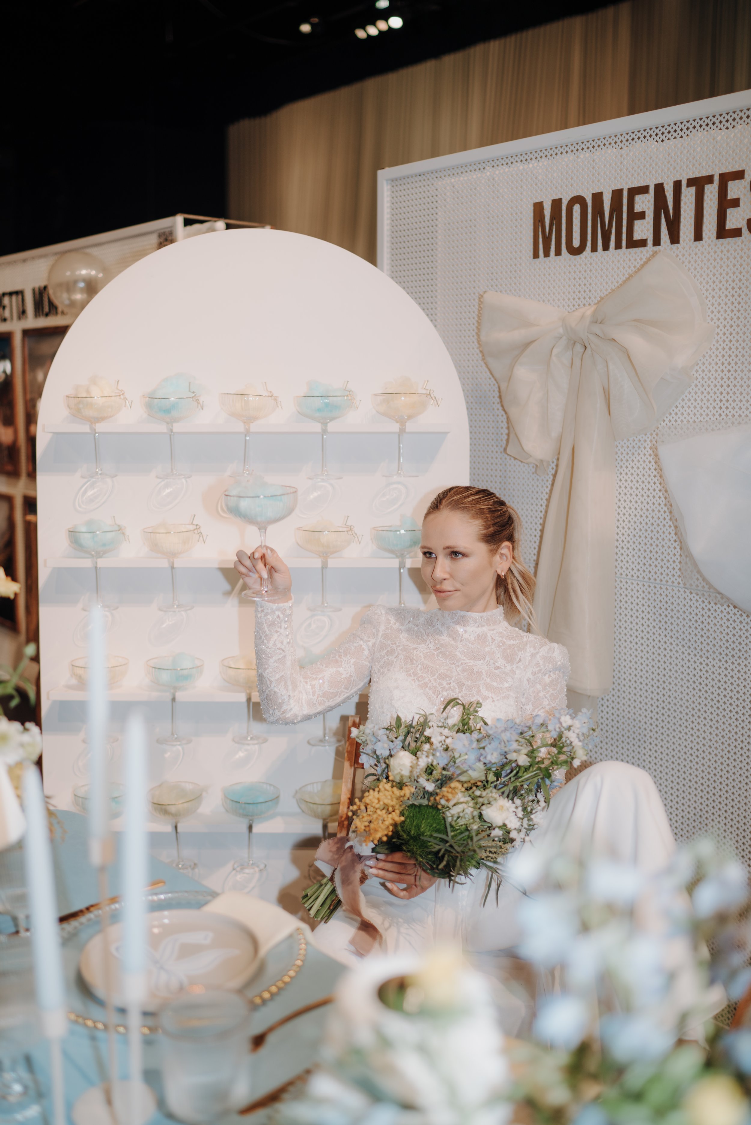 Eine junge Frau mit Hochzeitskleid sitzt an einem festlich gedeckten Tisch mit Blumen und hält einen Blumenstrauß, während sie im Hintergrund eine Tafel mit der Aufschrift „MOMENTES“ und einem großen weißen Schleifen-Garnitur bewundert.