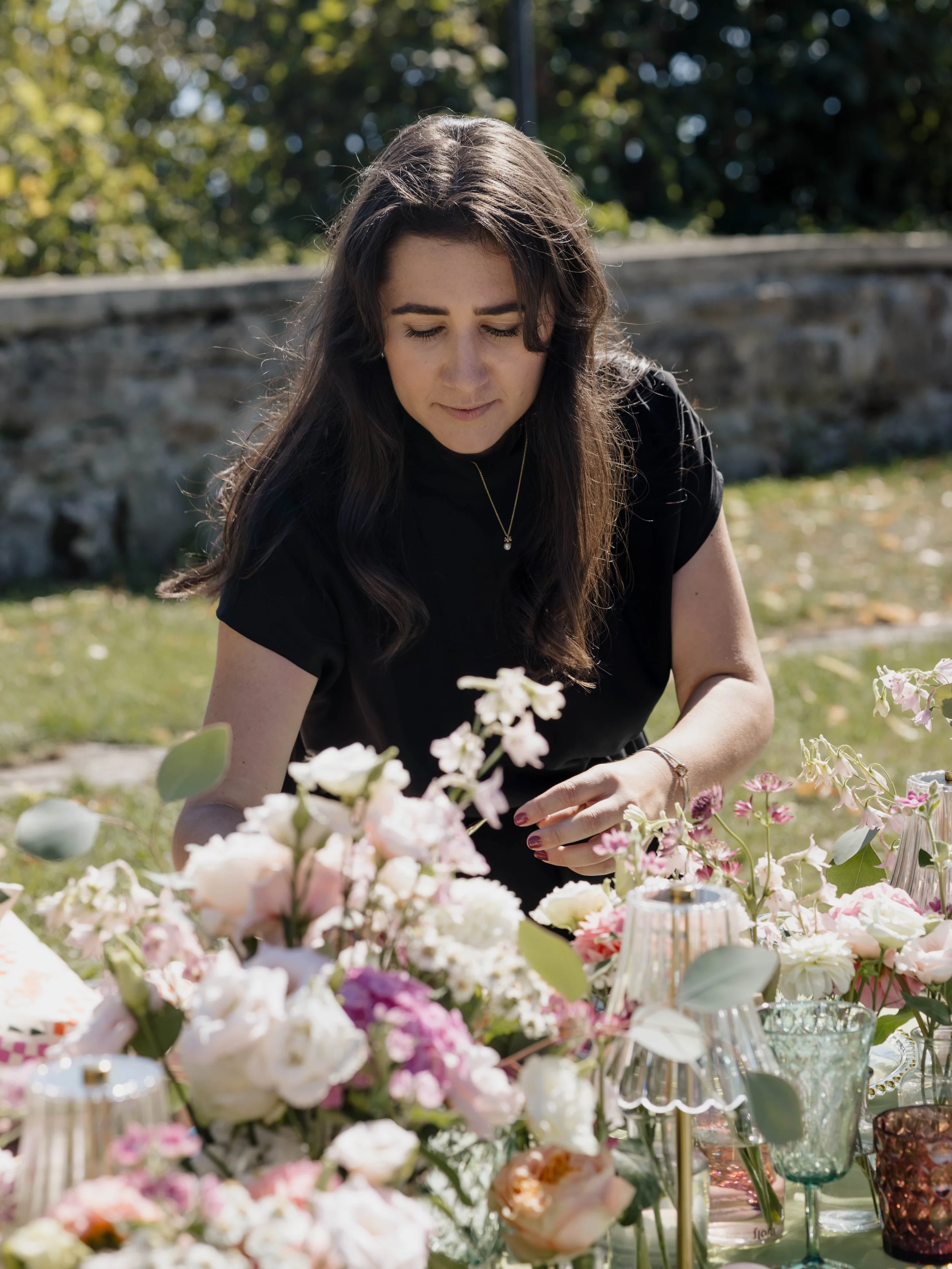 Eine Frau mit langen braunen Haaren und dunkler Kleidung, die bei schönem Wetter im Freien bei einer Dekoration mit vielen Blumen sitzt.