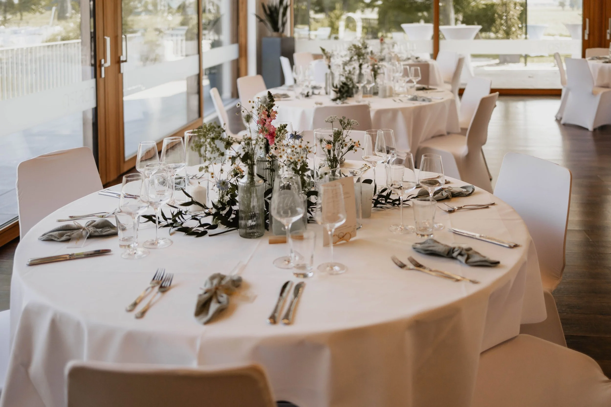 Ein rund gedeckter Tisch für eine Feier, mit weißen Tischdecken, Gläsern, Besteck, Stoffservietten und Blumendekoration in Vasen, in einem hellen Raum mit großen Fenstern.
