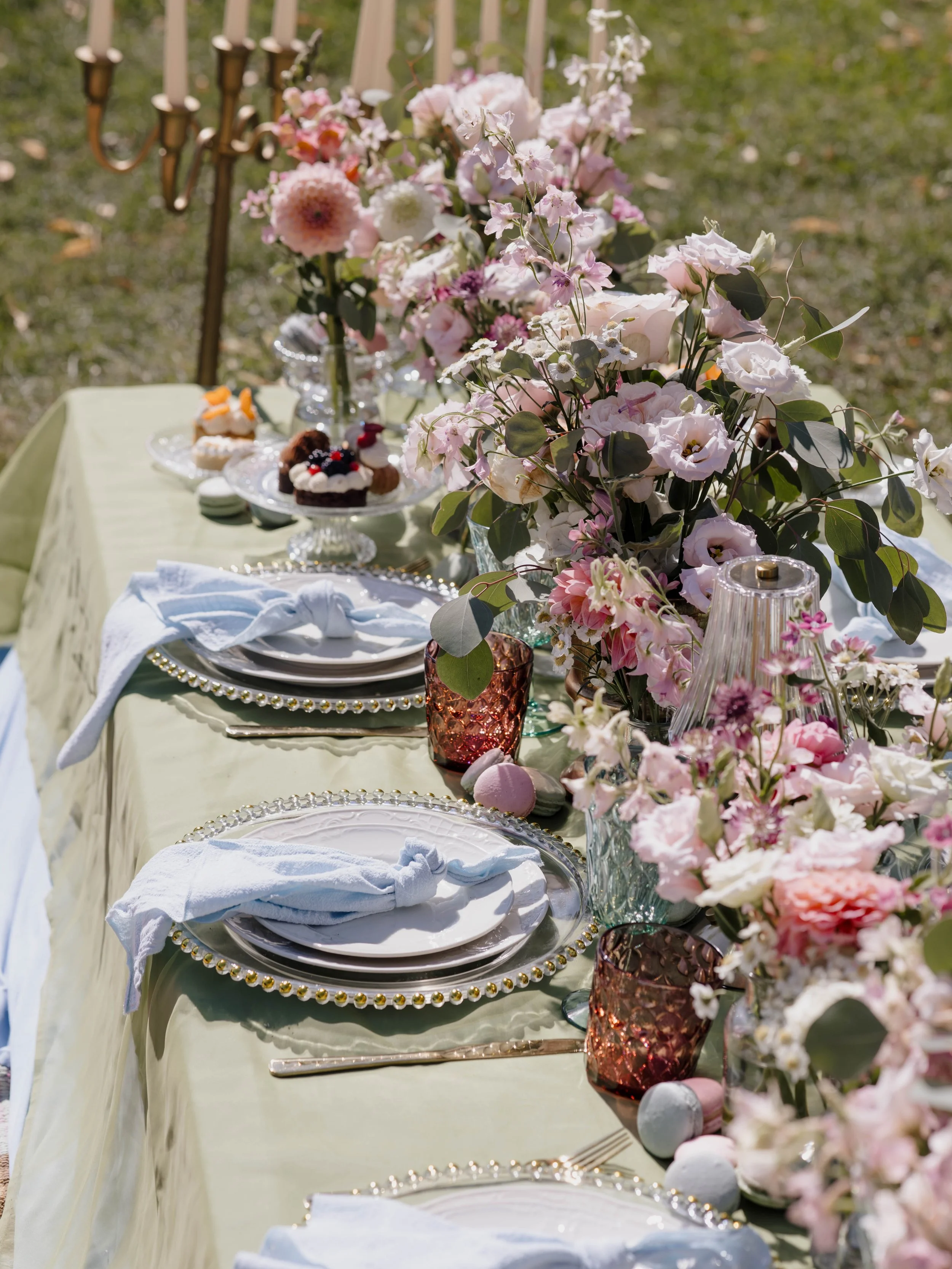 Tischdekoration mit Blumen, Geschirr und Desserts bei einer Feier im Außenbereich.