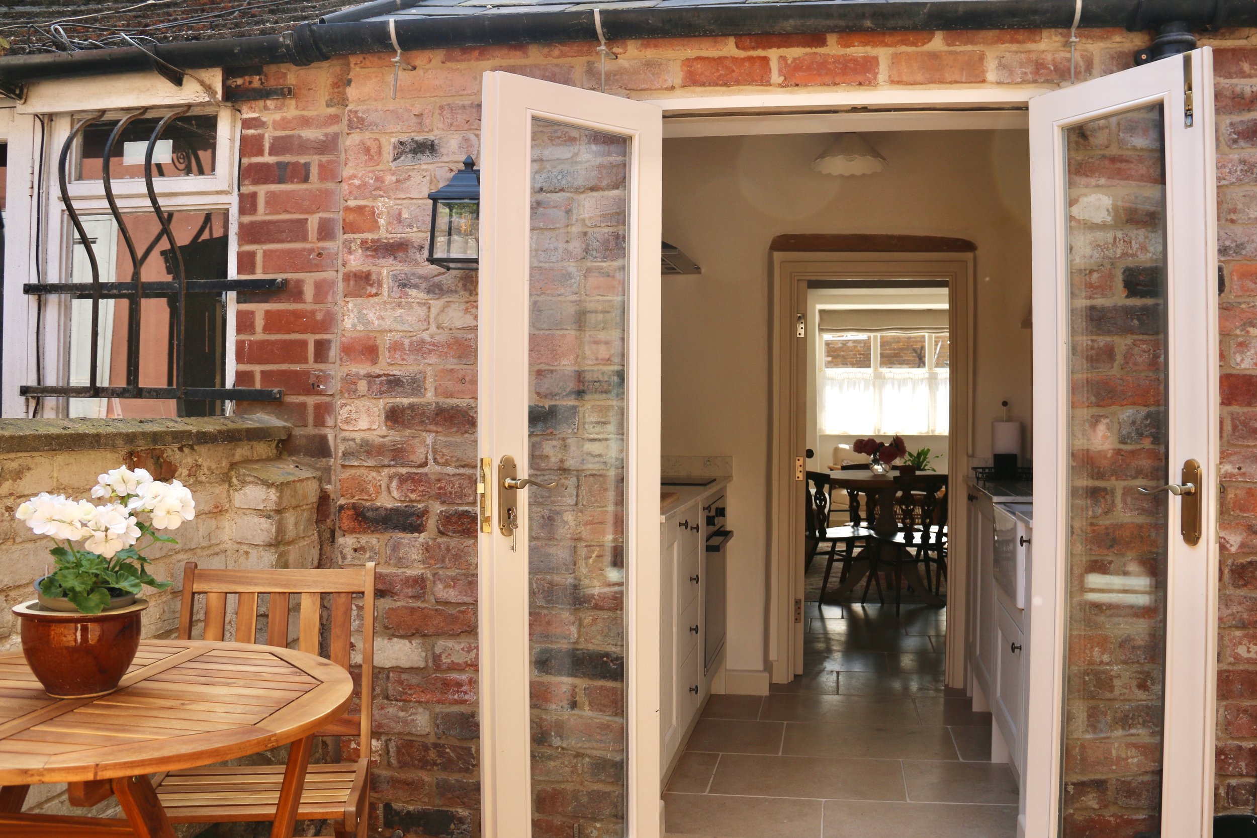 Uppingham Townhouse imagery of open white double doors leading from an outdoor brick patio into a cozy kitchen and dining area with a window and a small round dining table with flowers inside.