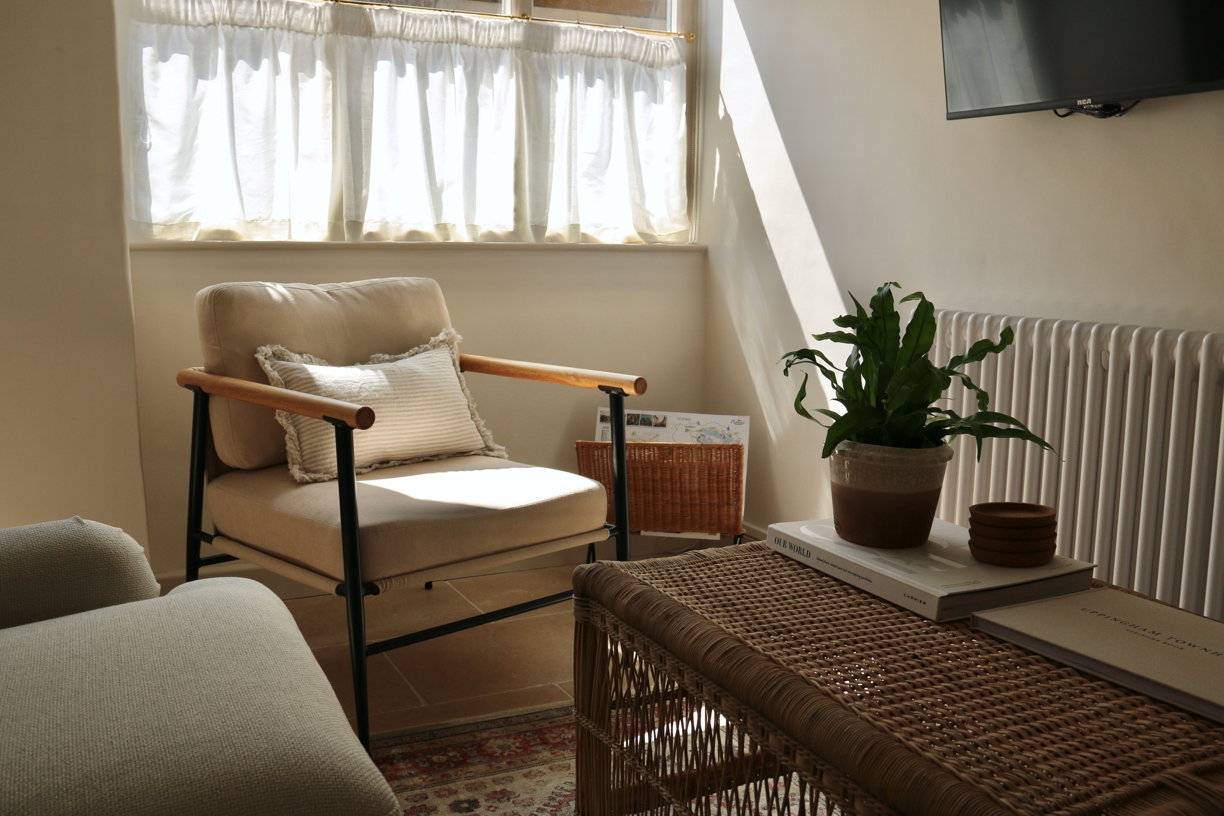 Uppingham Townhouse imagery of a cozy living room corner with sunlight streaming through cream curtains, a beige armchair with a pillow, a wicker side table with magazines and a potted plant, and a flat-screen TV on the wall.