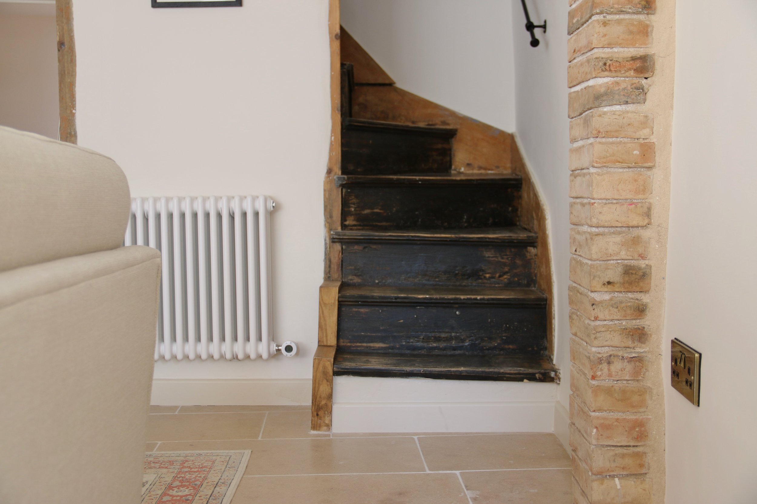 Uppingham Townhouse imagery of wooden stairs with black paint, next to a beige wall and brick pillar, with a white radiator and electrical outlet nearby.