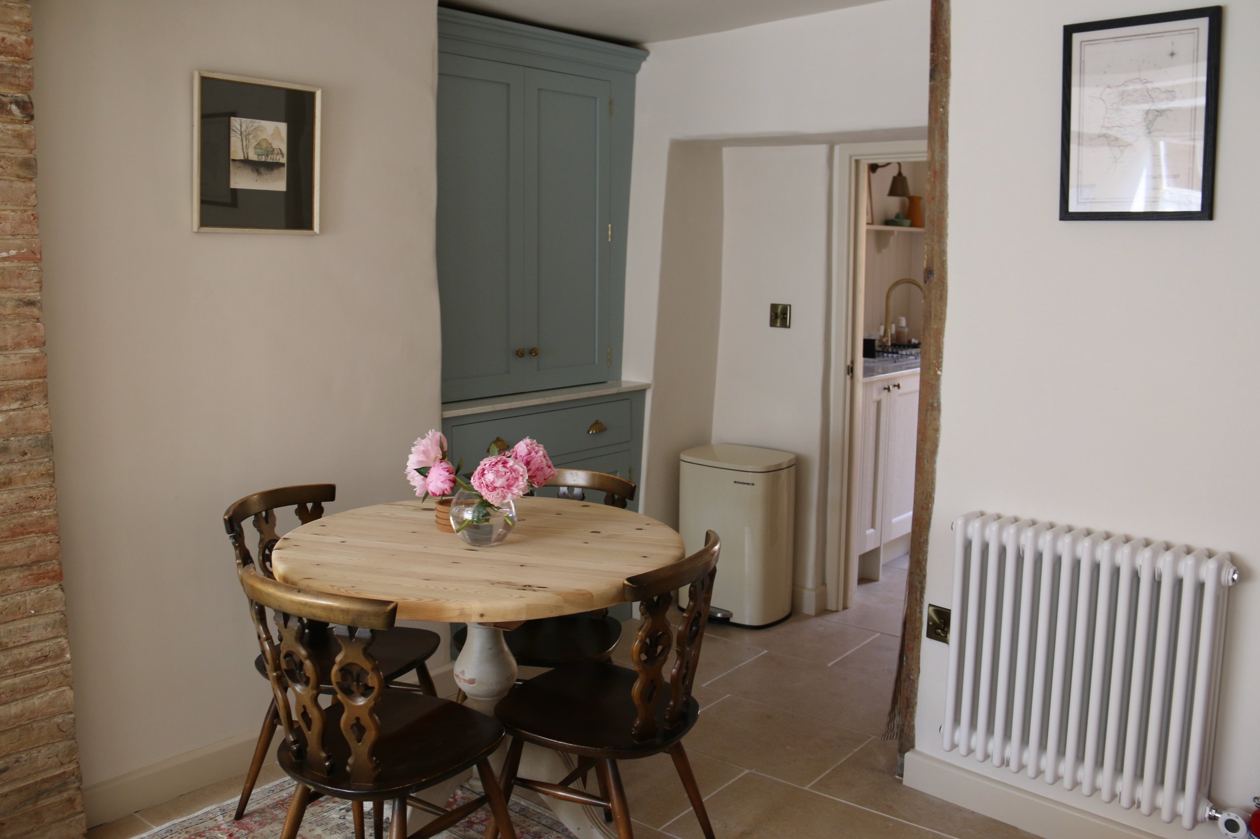 Uppingham Townhouse imagery of kitchen and dining area with a round wooden table, four wooden chairs, a vase with pink flowers, a grey cabinet, a beige trash bin, a white radiator, and framed artwork on the walls
