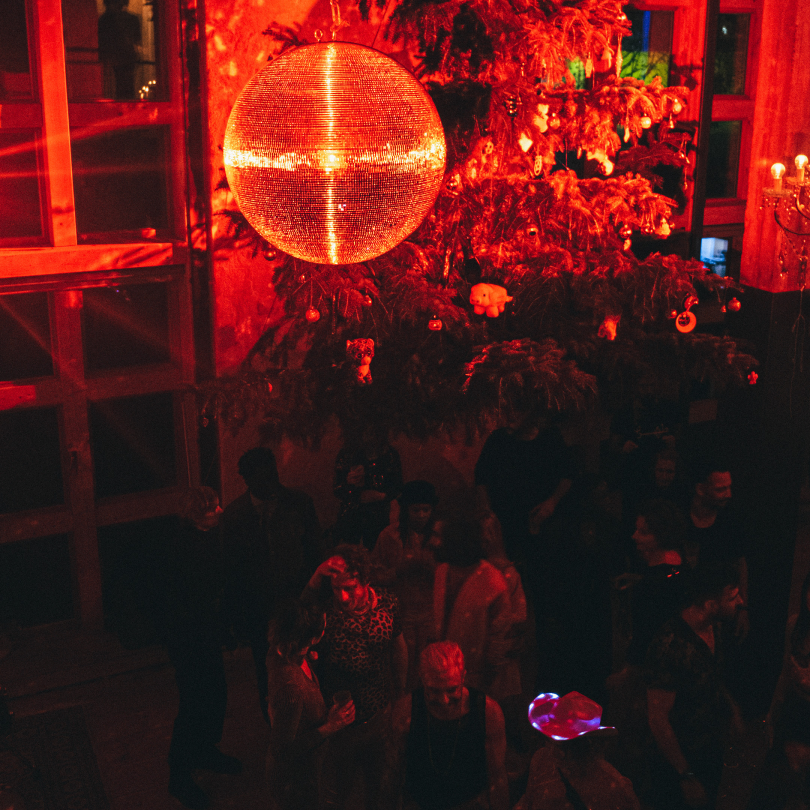 People socializing at a party with a decorated Christmas tree and a large disco ball illuminated in red lights.