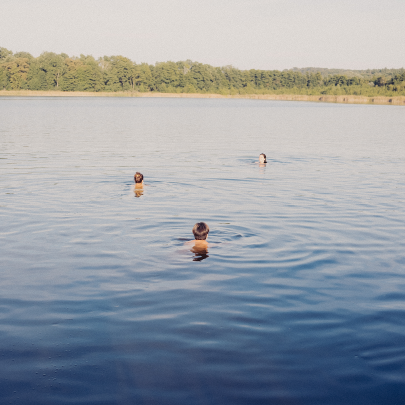 Three children swimming in a lake with a tree-lined shoreline in the background, under clear sky.