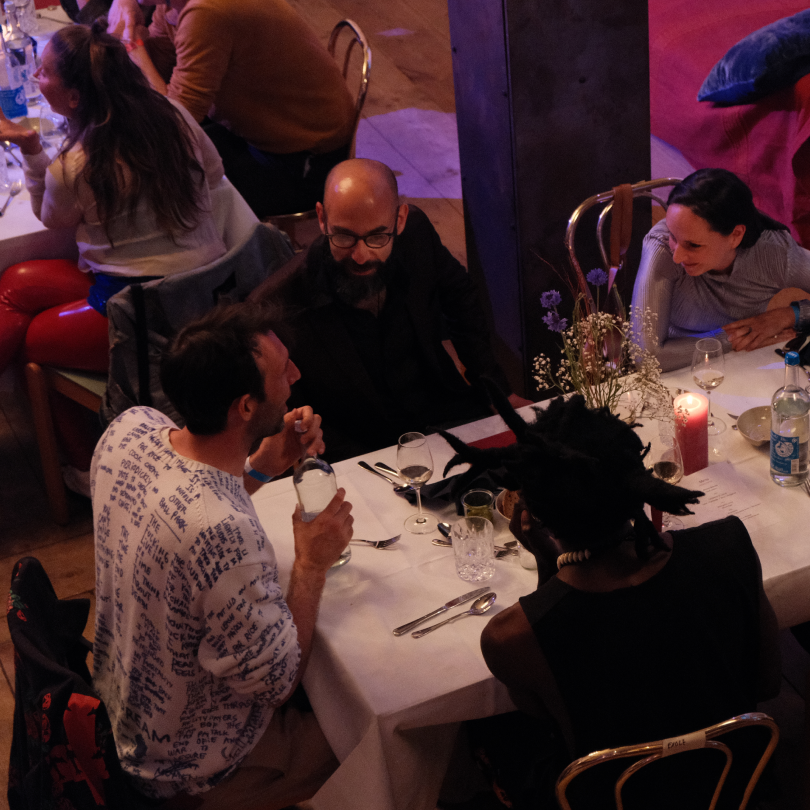 People sitting around a dining table having a conversation in a dimly lit restaurant, with plates, glasses, a candle, and flowers on the table.