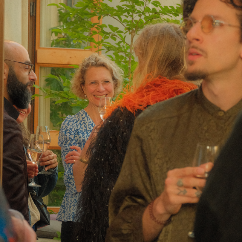 Group of people at a social gathering, holding wine glasses, engaging in conversation, with a woman smiling in the background near a window with greenery outside.