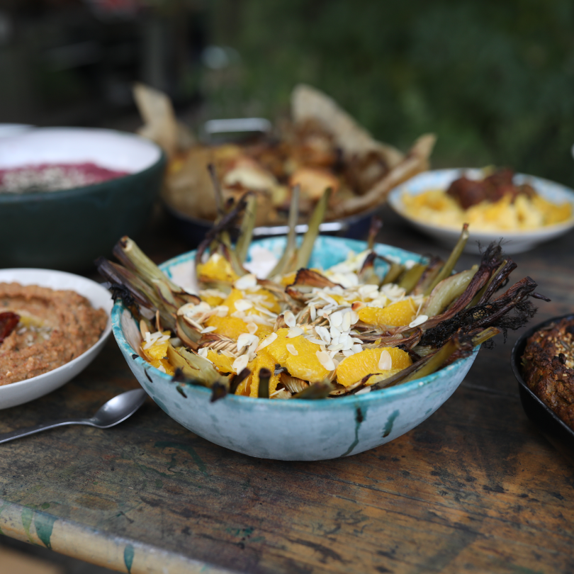 A rustic outdoor table with a large bowl of grilled artichokes topped with orange slices and almond flakes, surrounded by various dishes including dips and roasted vegetables.
