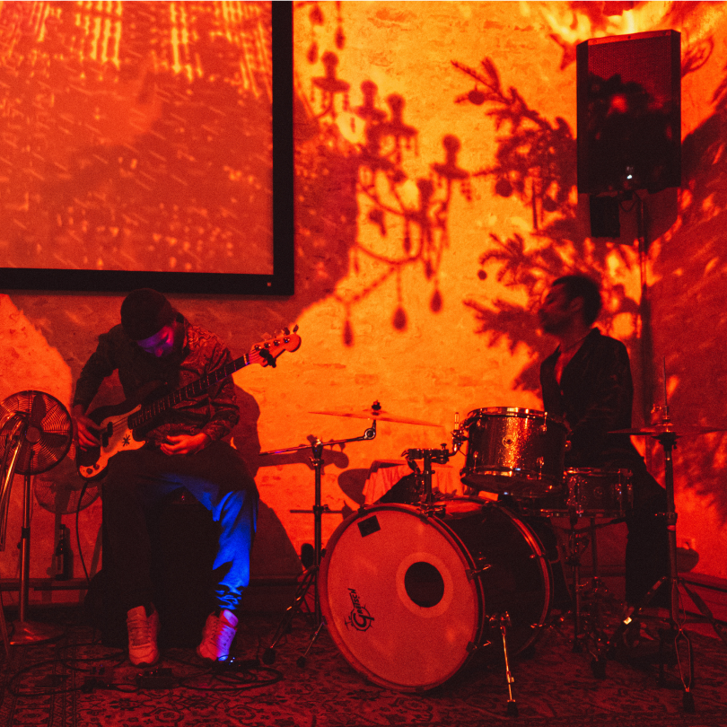 Two musicians performing in a dimly lit room with orange and red lighting. One is playing an electric guitar and the other is on drums. Shadows of tree branches are projected onto the wall behind them.