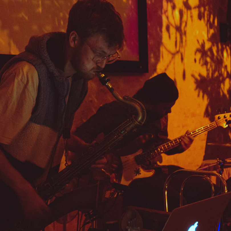 Two musicians playing instruments in a dimly lit room, one with a saxophone and the other with a bass guitar, with a warm, orange-hued background.