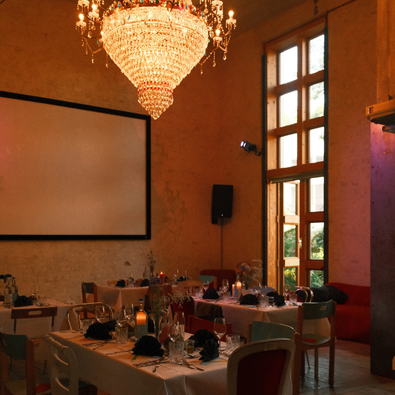 Elegant dining room with a large chandelier, decorated tables with black napkins, wine glasses, and candles, and tall wooden windows revealing a view outside.