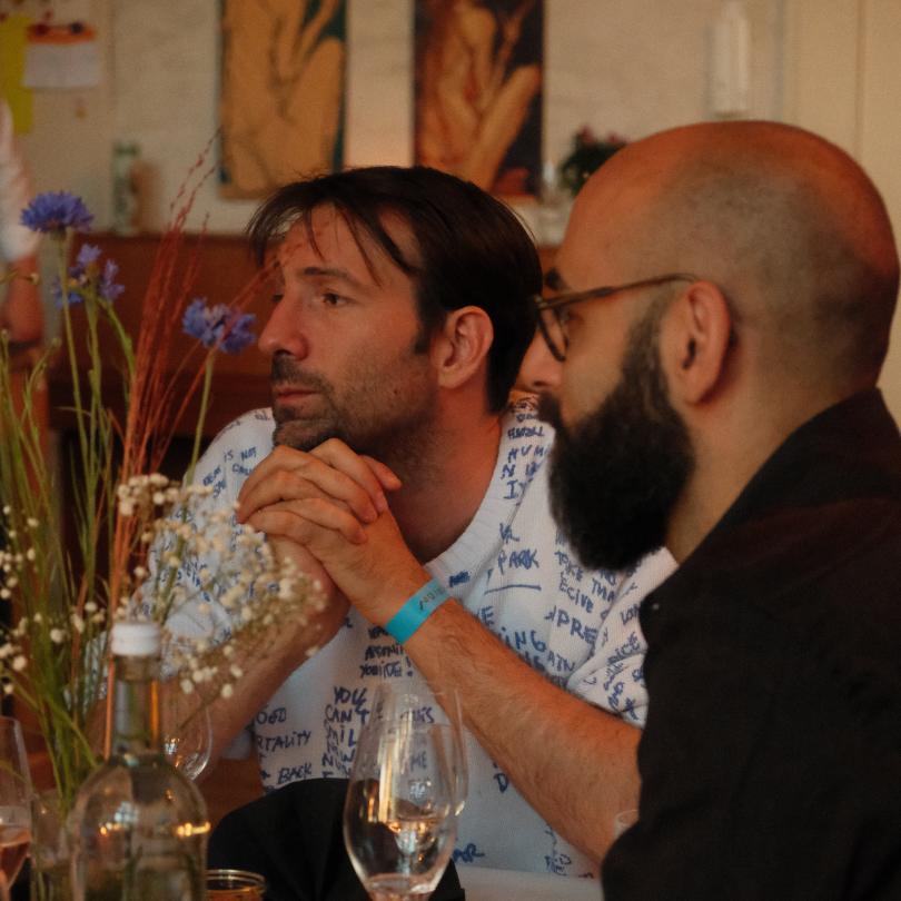 Two men sitting at a table, one with hands clasped and the other with a beard and glasses, in a cozy restaurant setting with flowers and artwork in the background.