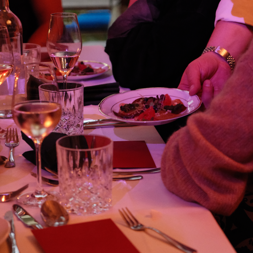 People holding a plate of food at a formal dinner table with glasses of wine and elegant tableware.