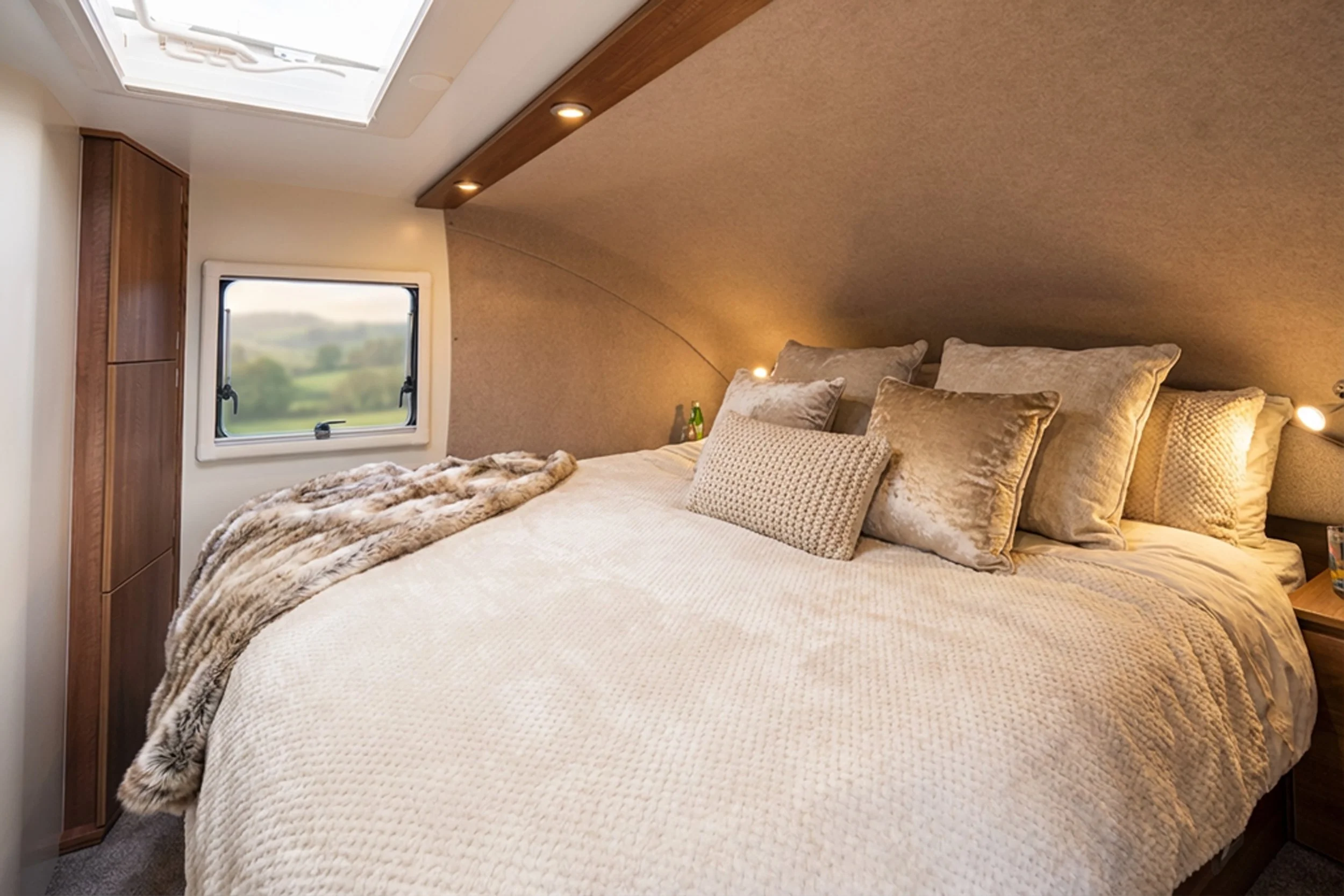 Interior of a small RV showing a bed with pillows and blankets, an alarm clock, and a nautical decoration. Adjacent is a compact shower area with a handheld showerhead.