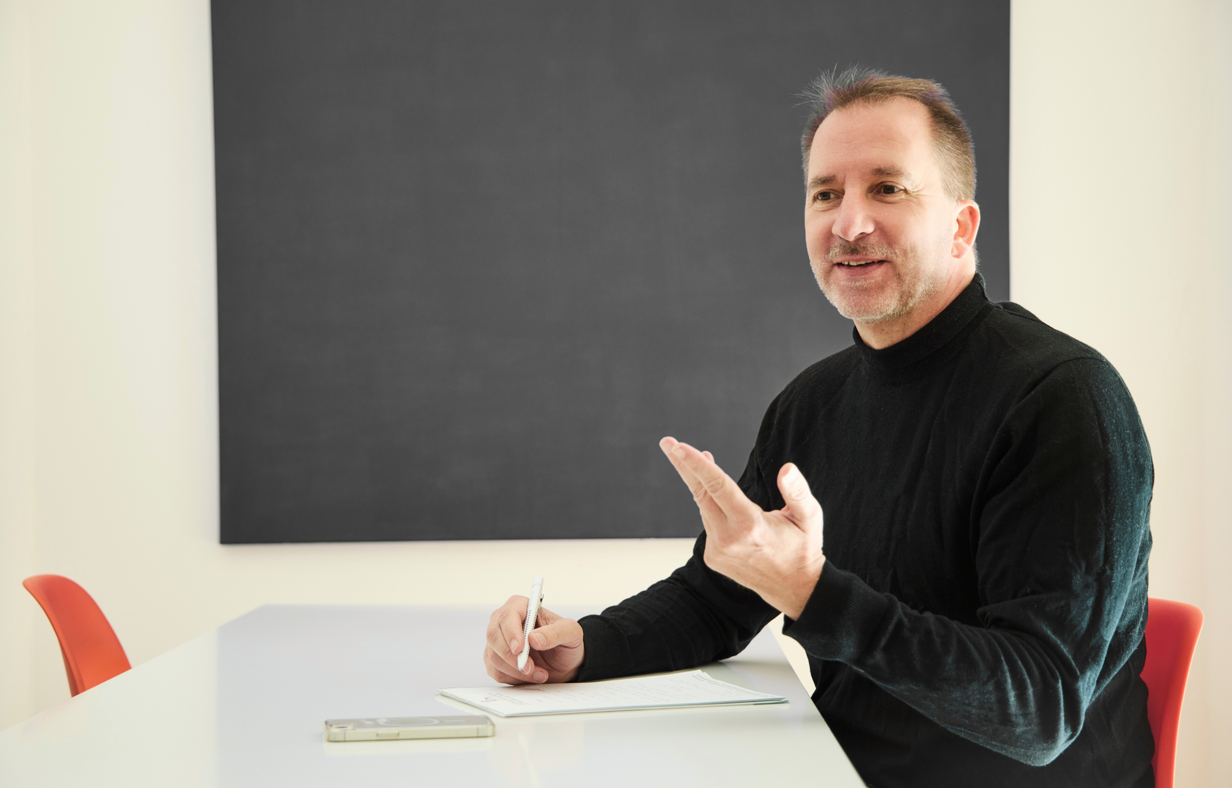 Andreas Berglitz sitzt an einem weißen Tisch, hält einen Stift in der Hand und schaut nach rechts, während er spricht. Im Hintergrund eine große schwarze Tafel.