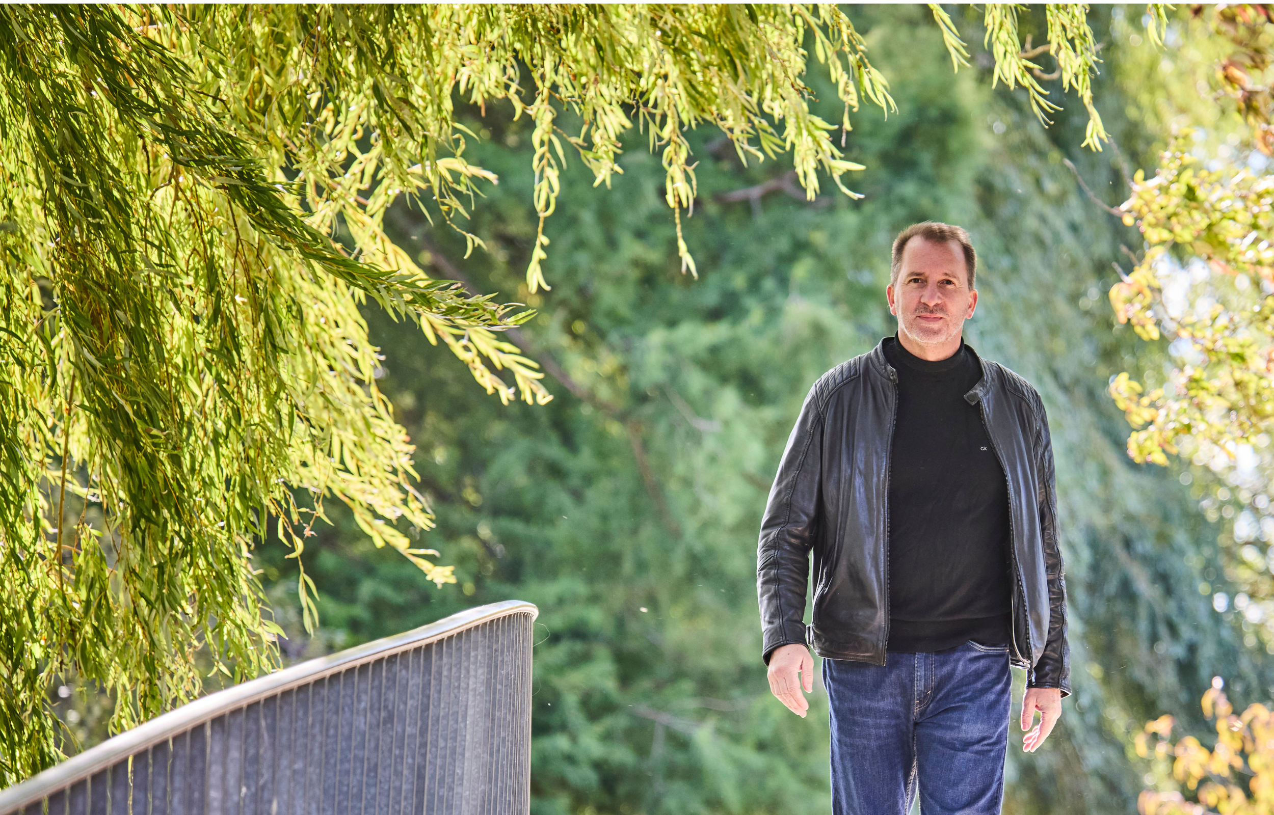 Andreas Berglitz geht in dunkler Lederjacke und blauen Jeans über eine Brücke durch einen parkähnlichen Bereich mit grünen Bäumen.