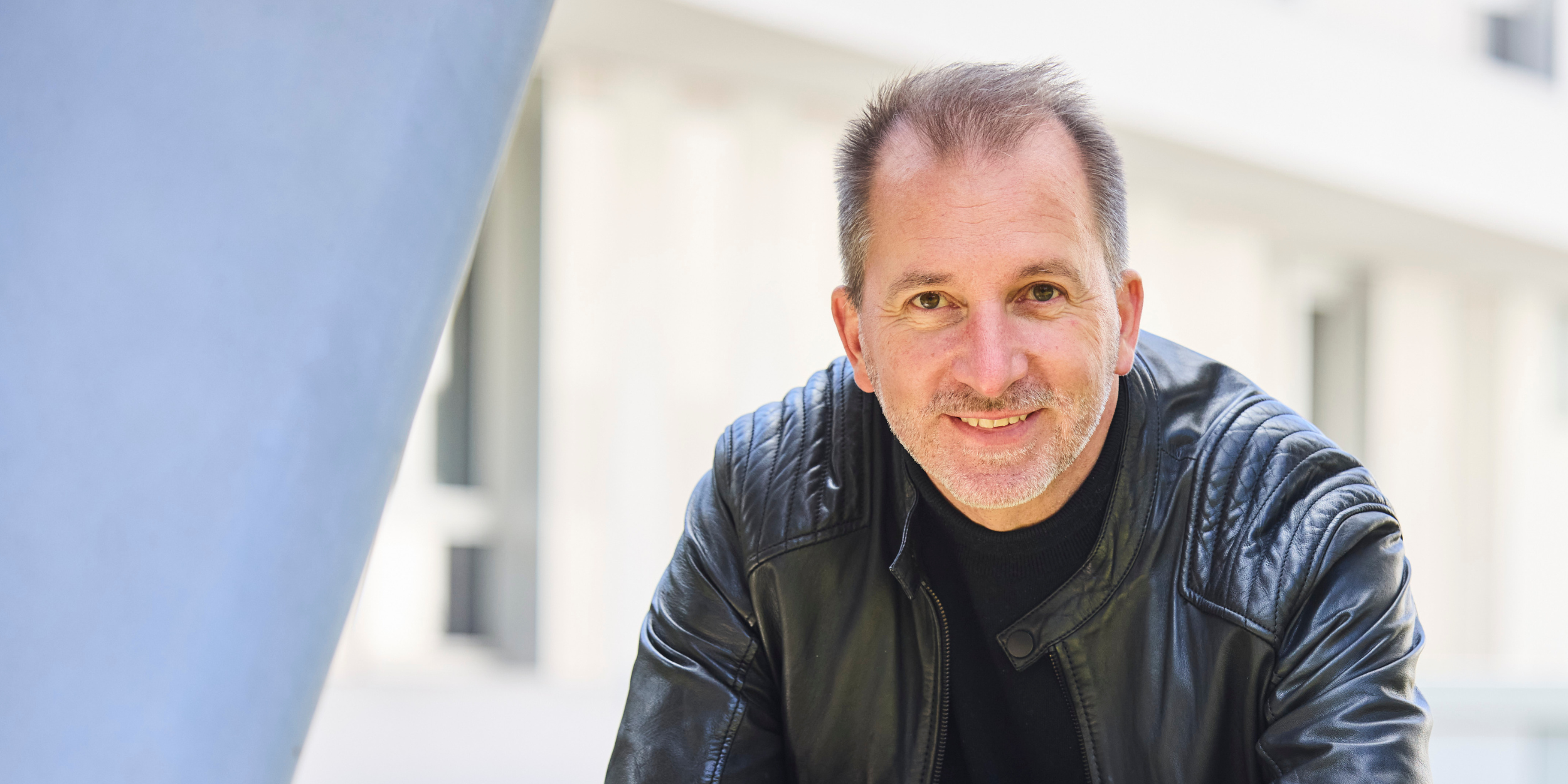 Ein Mann mit kurzen, grauen Haaren und einem lockeren Lächeln, trägt eine schwarze Lederjacke, steht vor einem hellen Hintergrund im Freien.
