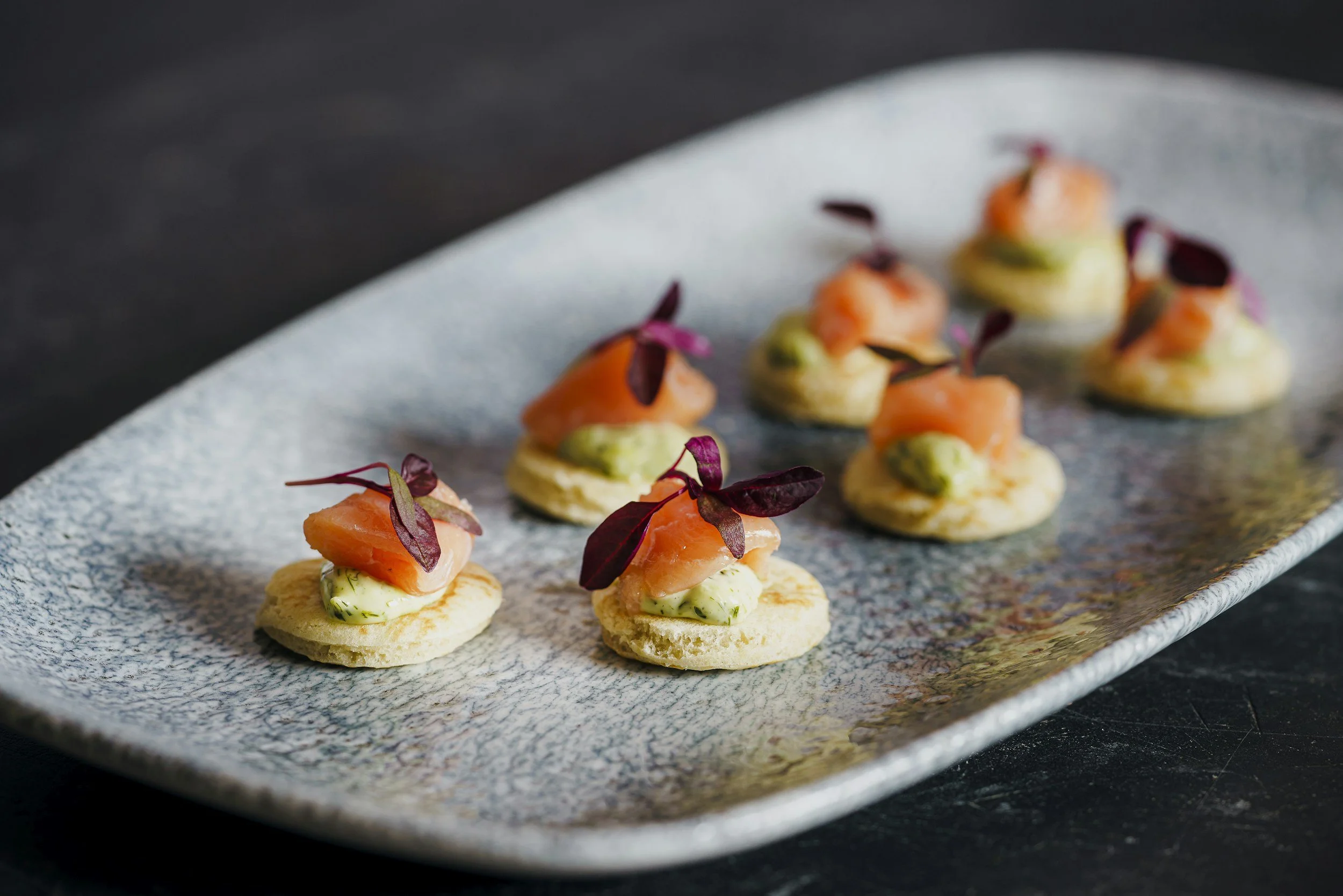 Smoked salmon canapé with a dollop of cream and purple microgreens on a textured rectangular plate.