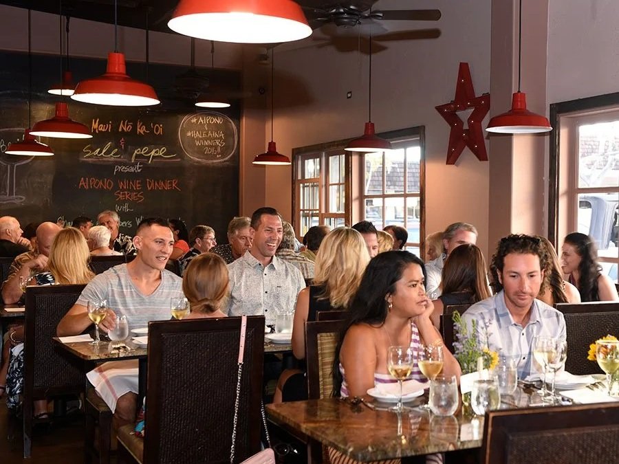 A lively restaurant filled with people dining and socializing, with red pendant lights, large windows, and a chalkboard wall with handwritten menu and announcements.
