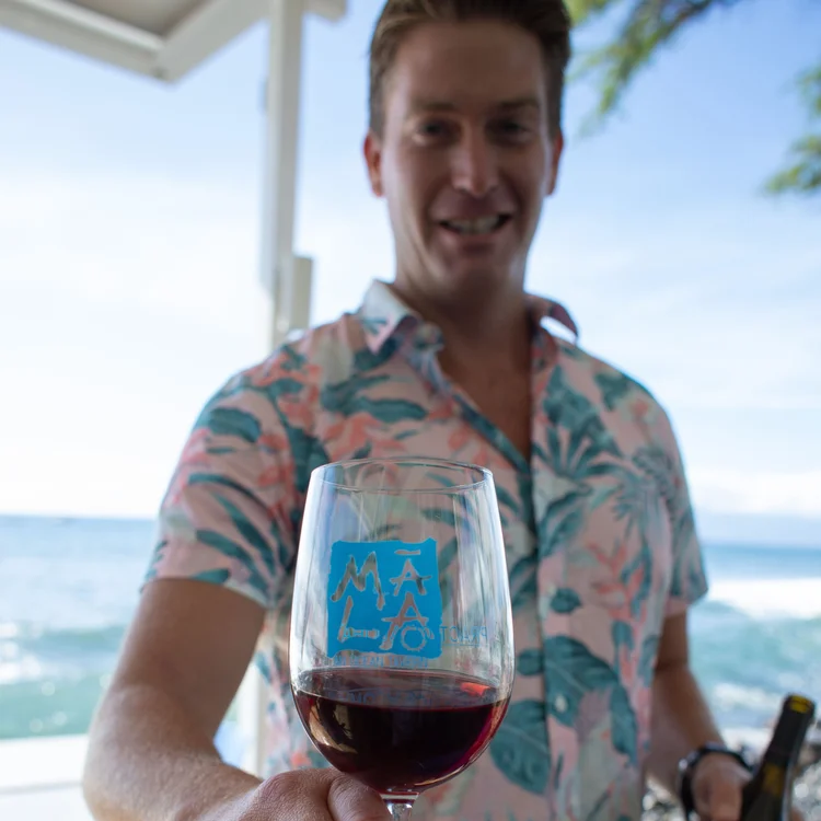 Man in a floral shirt at the beach holding a glass of red wine with a blue logo on it.