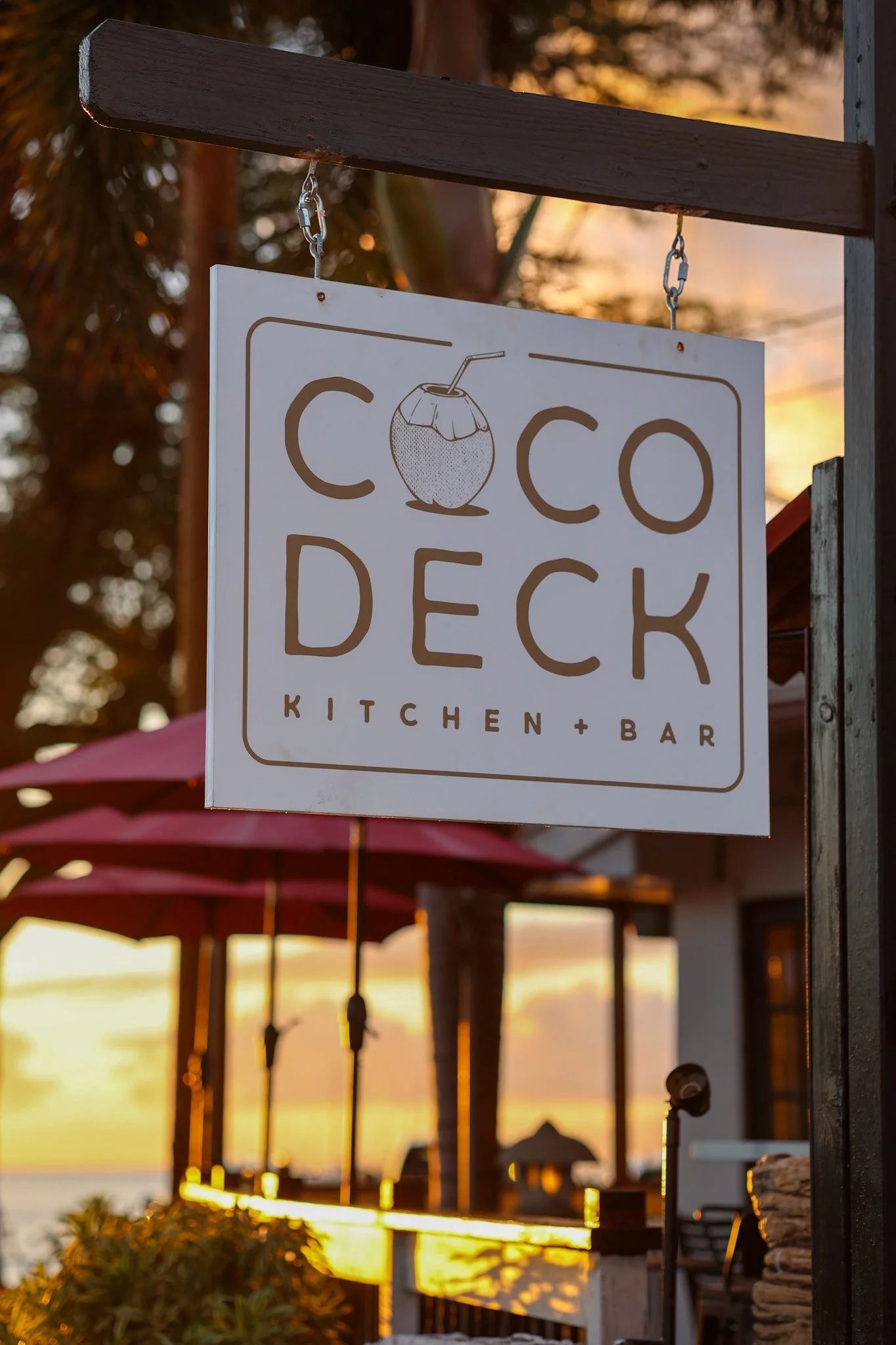 Sign for Coco Deck Kitchen and Bar hanging outdoors at sunset with a view of the beach and umbrellas.