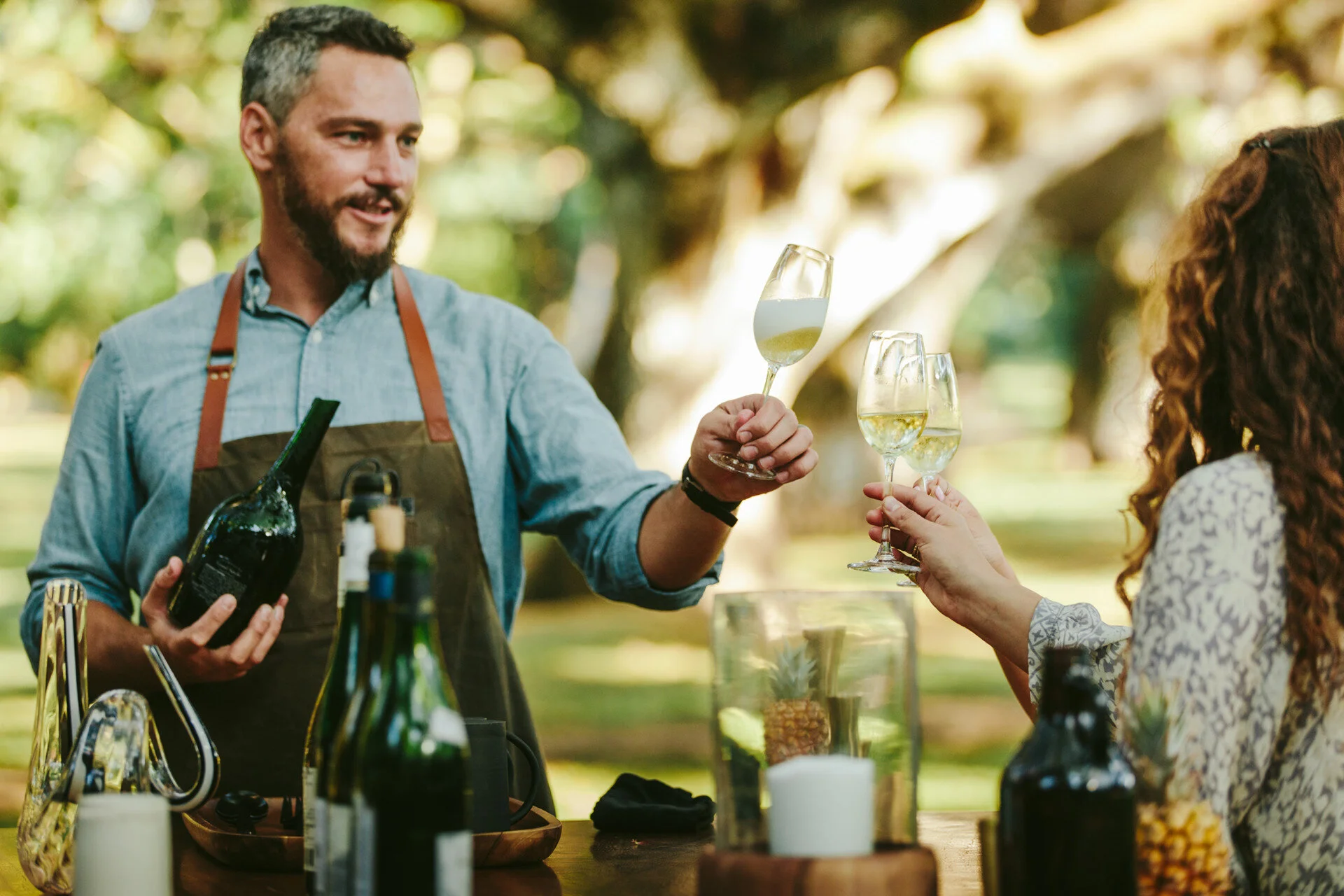 A man with a beard and blue shirt wearing an apron is brewing wine or another alcoholic beverage outdoors. He is holding a wine bottle and a wine glass, handing it to a woman with curly hair who is also holding a glass. The scene takes place in a park or garden with trees and greenery in the background.