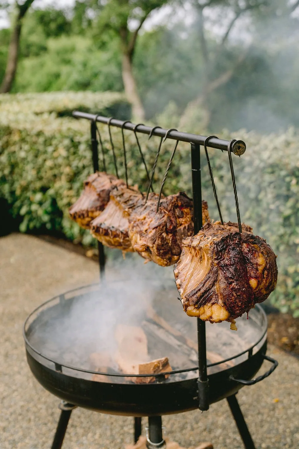 Four large pieces of meat hanging on a metal rack over an open grill, with smoke rising in an outdoor setting with trees and greenery in the background.