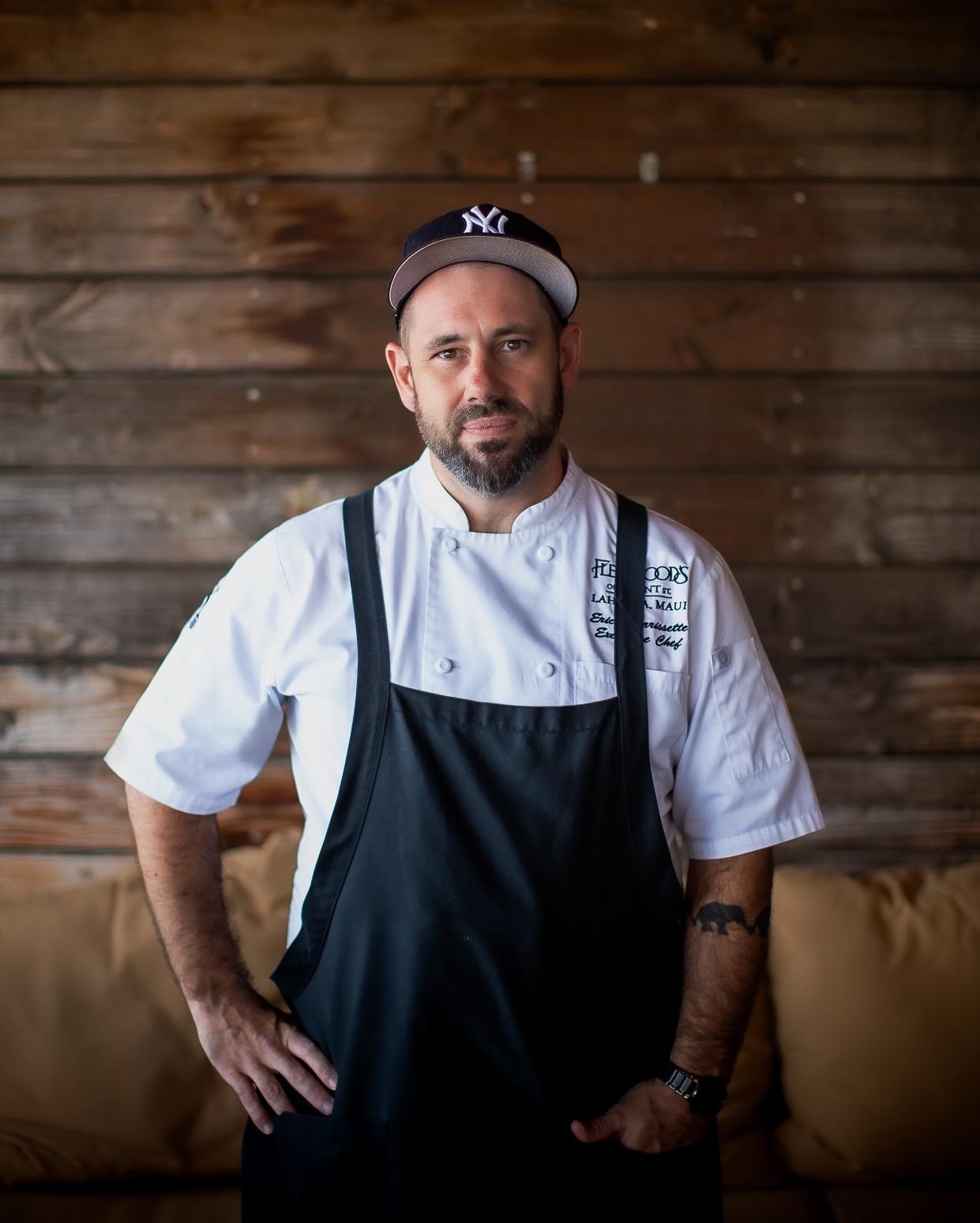 A male chef with a beard, wearing a white chef's coat, black apron, a watch, and a black New York Yankees cap, standing indoors against a wooden wall.