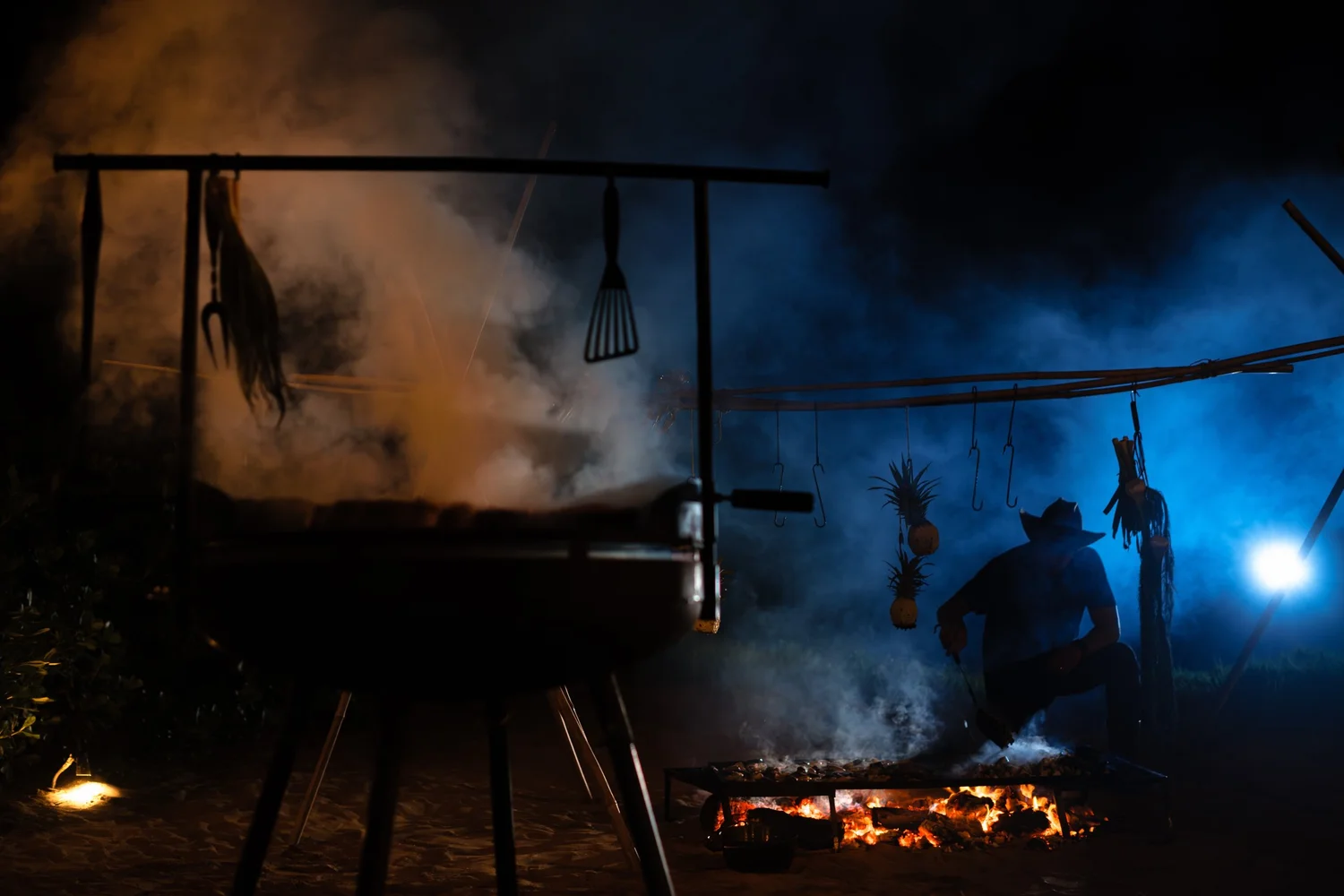 A person crouching near an outdoor fire at night, with hanging pineapple decorations, garden tools, and smoke illuminated by a bright blue light.