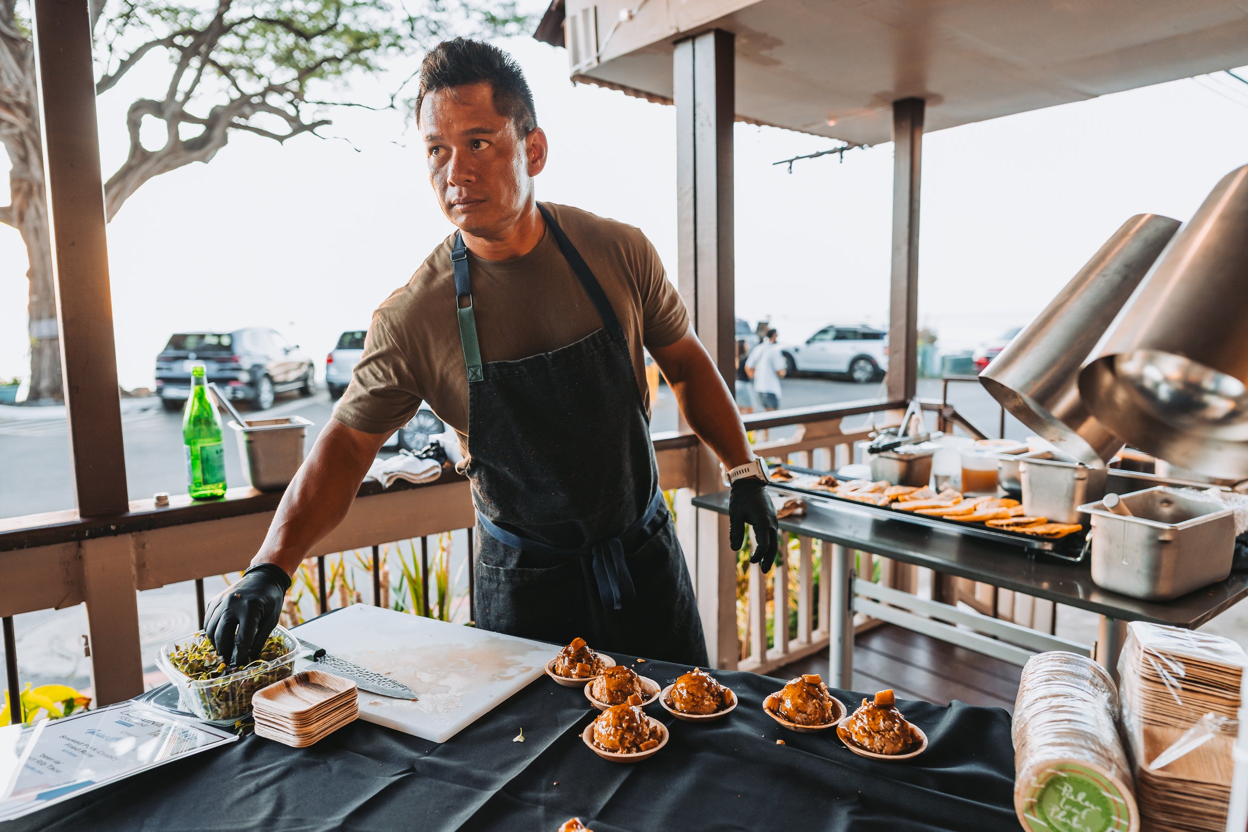 A man wearing a black apron and gloves preparing food at a buffet table outdoors, with plates of food, a green bottle, and cooking equipment nearby.