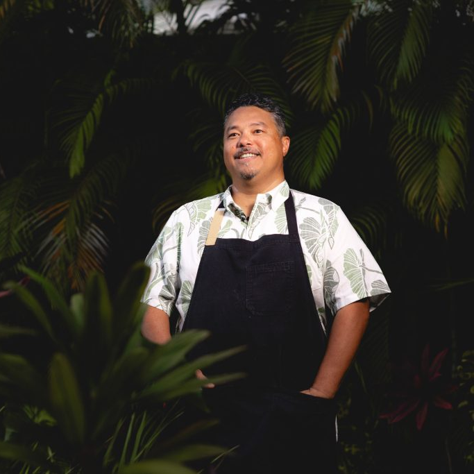 Man wearing a patterned short-sleeve shirt and black apron, standing outdoors amid lush green tropical plants, smiling.