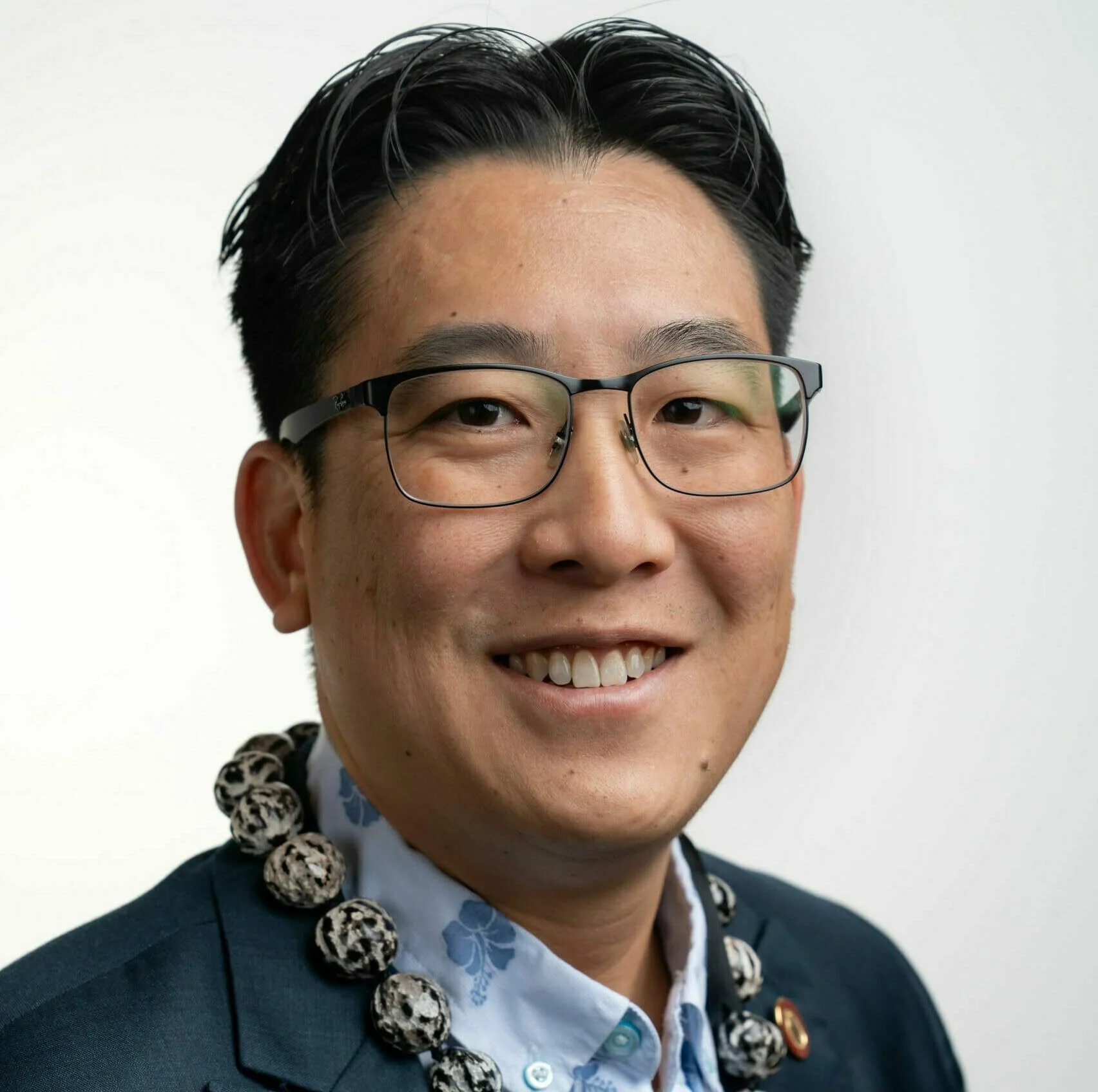 A man wearing glasses, a blue blazer, a blue patterned shirt, and a necklace with large black and white beads, standing against a plain white background and smiling.