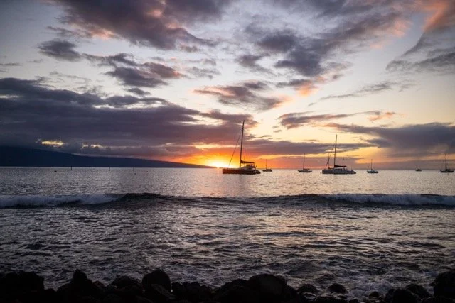 Sunset over the ocean with several sailboats anchored near the shore and a rocky beach in the foreground. View from Coco Deck