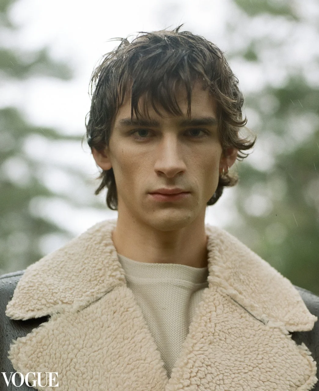 Editorial Porträt in natürlichem, diffusem Outdoor-Licht: ein intimer Close-up mit filmischem Grain, ruhiger Spannung im Ausdruck und kühlen, gedämpften Grüntönen im Hintergrund. Der voluminöse Shearlingkragen setzt eine starke Textur gegen die glatt