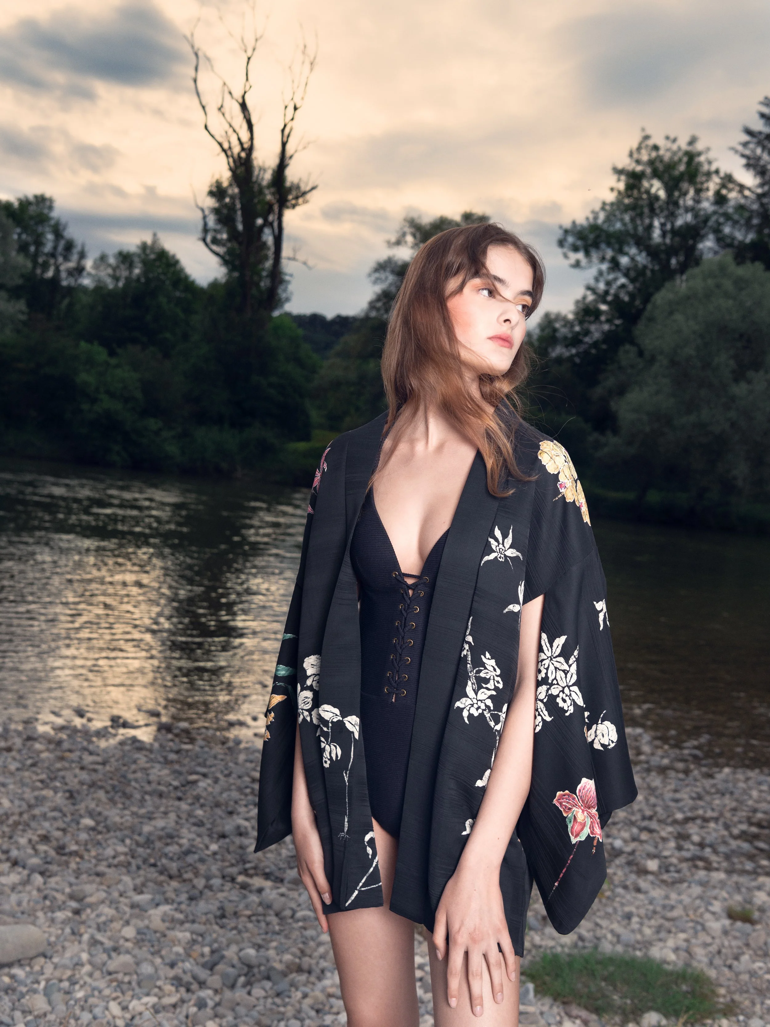 Eine junge Frau steht am Ufer eines ruhigen Flusses, umgeben von dichter Natur. Der Himmel ist dramatisch bewölkt, das Licht sanft und diffus. Sie trägt einen tief ausgeschnittenen, geschnürten schwarzen Badeanzug, darüber einen bestickten Kimono mit