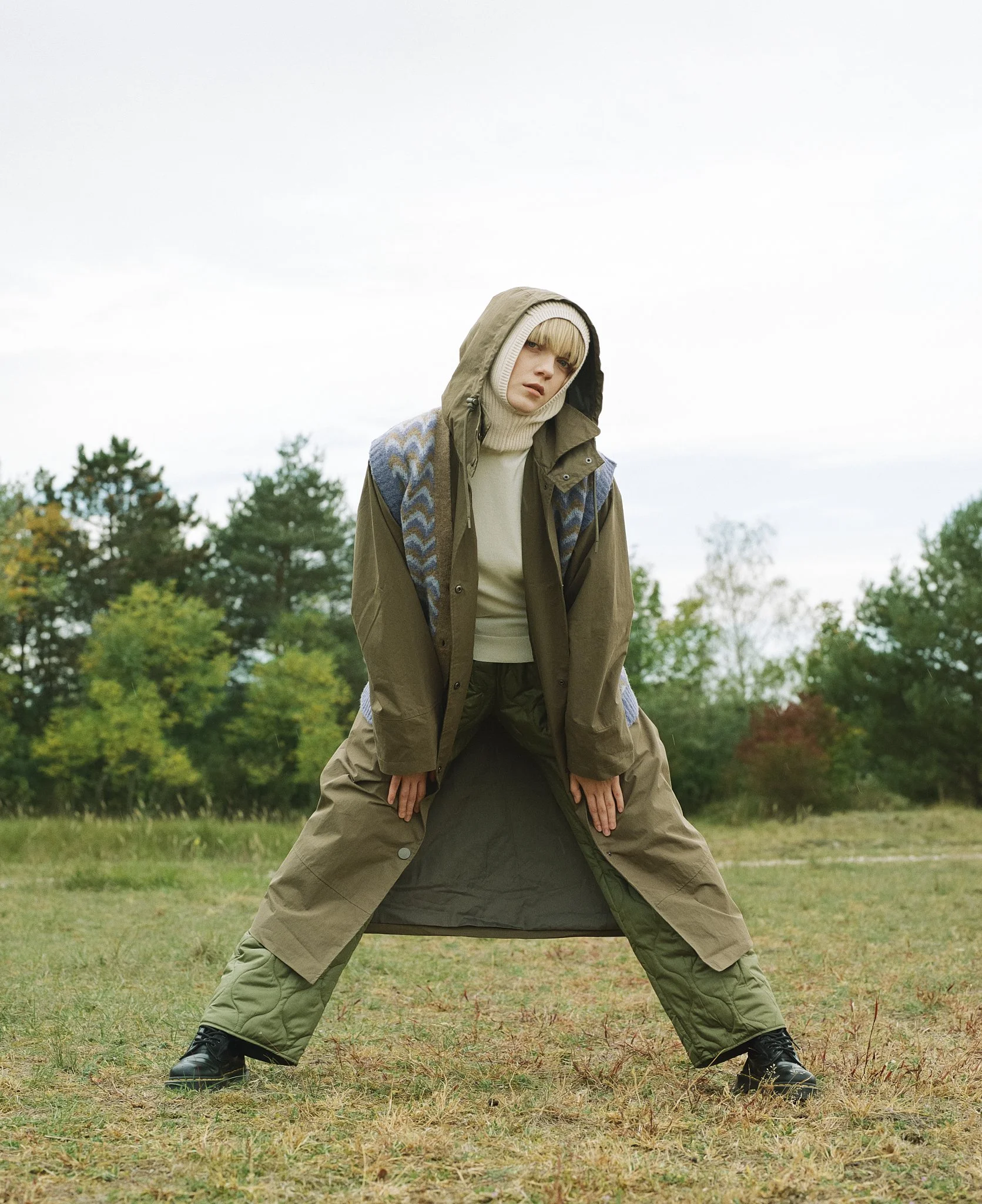 Model in khakifarbenem Mantel, Strickjacke und Mütze, in dynamischer Pose mit natürlichem Make-up vor natürlichem Hintergrund.