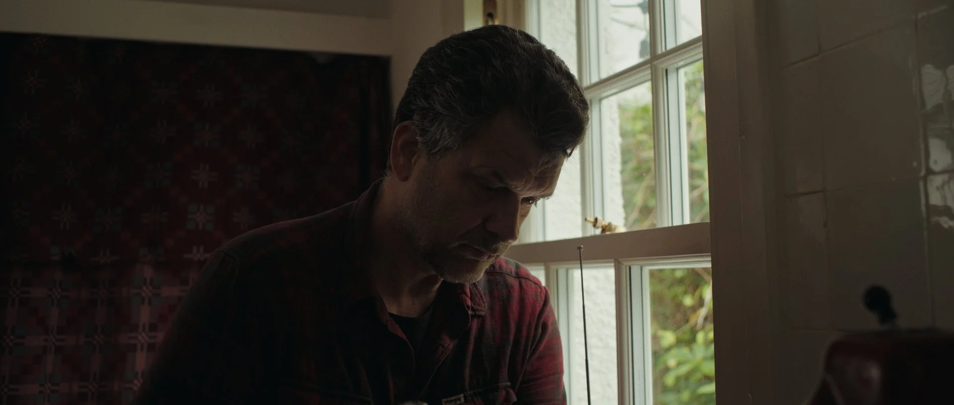 Wider shot of Jan-Jacob slicing bacon, the natural light wrapping around his silhouette