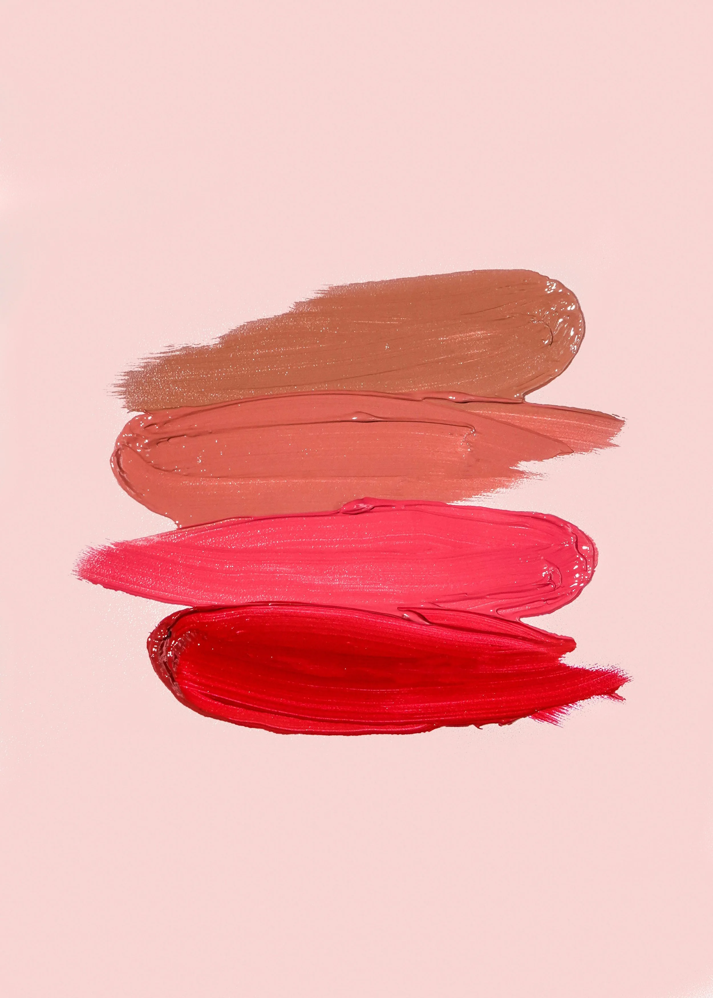 Quatre coups de pinceau de différentes couleurs de rouge à lèvres sur fond rose clair, allant du beige au rouge vif.