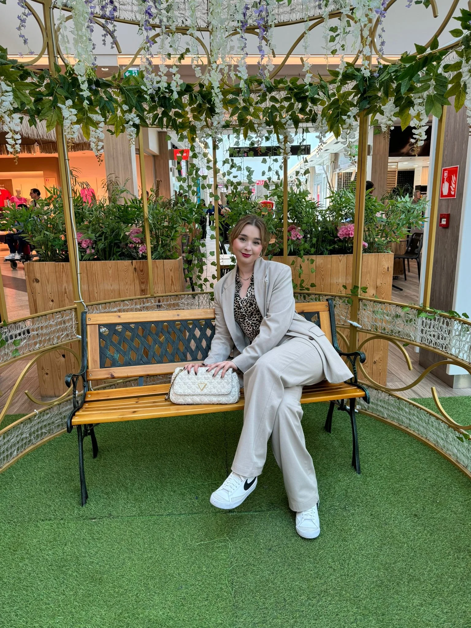 Jeune femme assise sur un banc en bois dans un espace décoré de plantes vertes et de fleurs, portant une tenue beige, des baskets Nike blanches avec le logo noir, une chemise léopard, et tenant un sac à main clair sur le banc.
