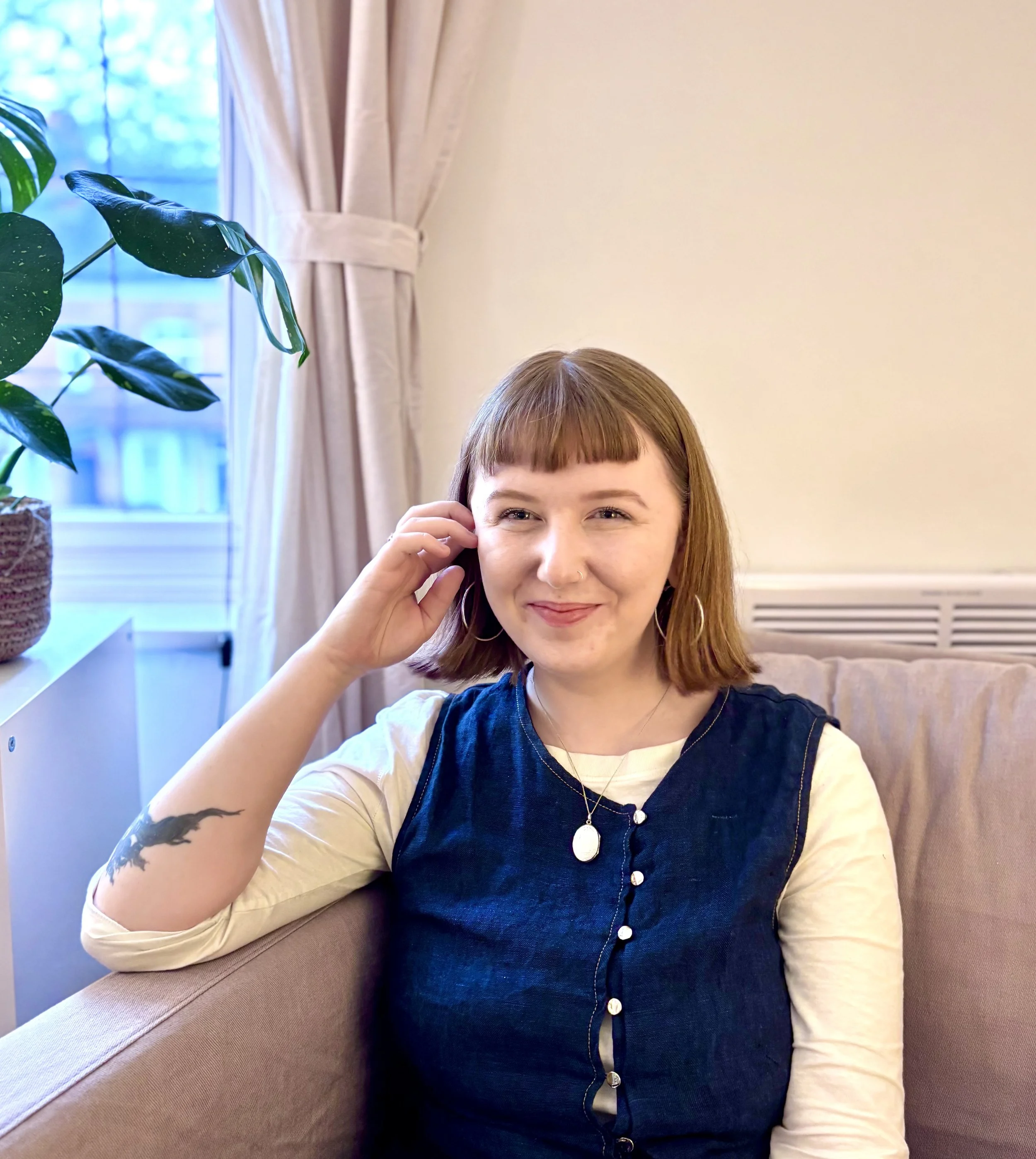 Team member Emma Buckley wearing a denim dress over a cream-colored long sleeve shirt, sitting on a beige couch with a smile, with a large green houseplant and a window with cream curtains in the background.