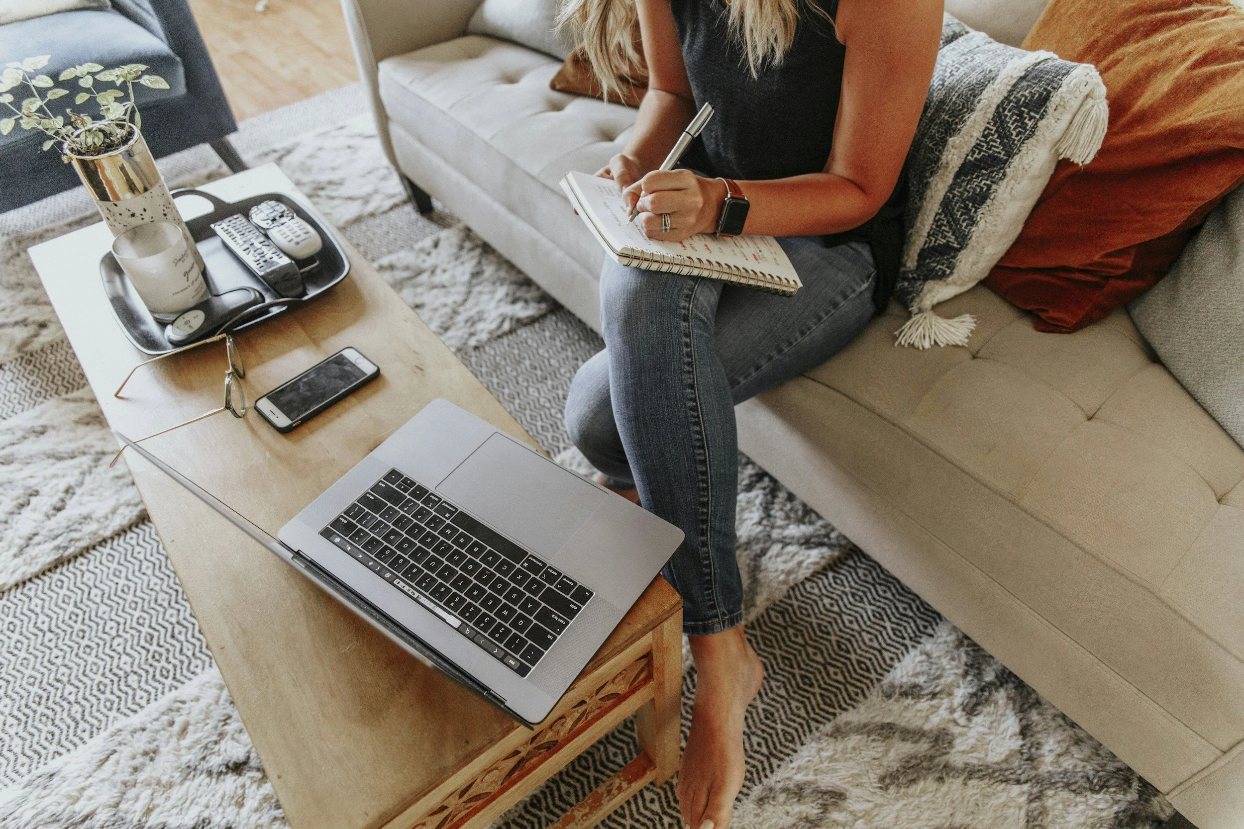 Woman sitting on a beige sofa, writing in a notebook, with a laptop, smartphone, glasses, and a tray with a plant and mugs on a wooden coffee table in front.