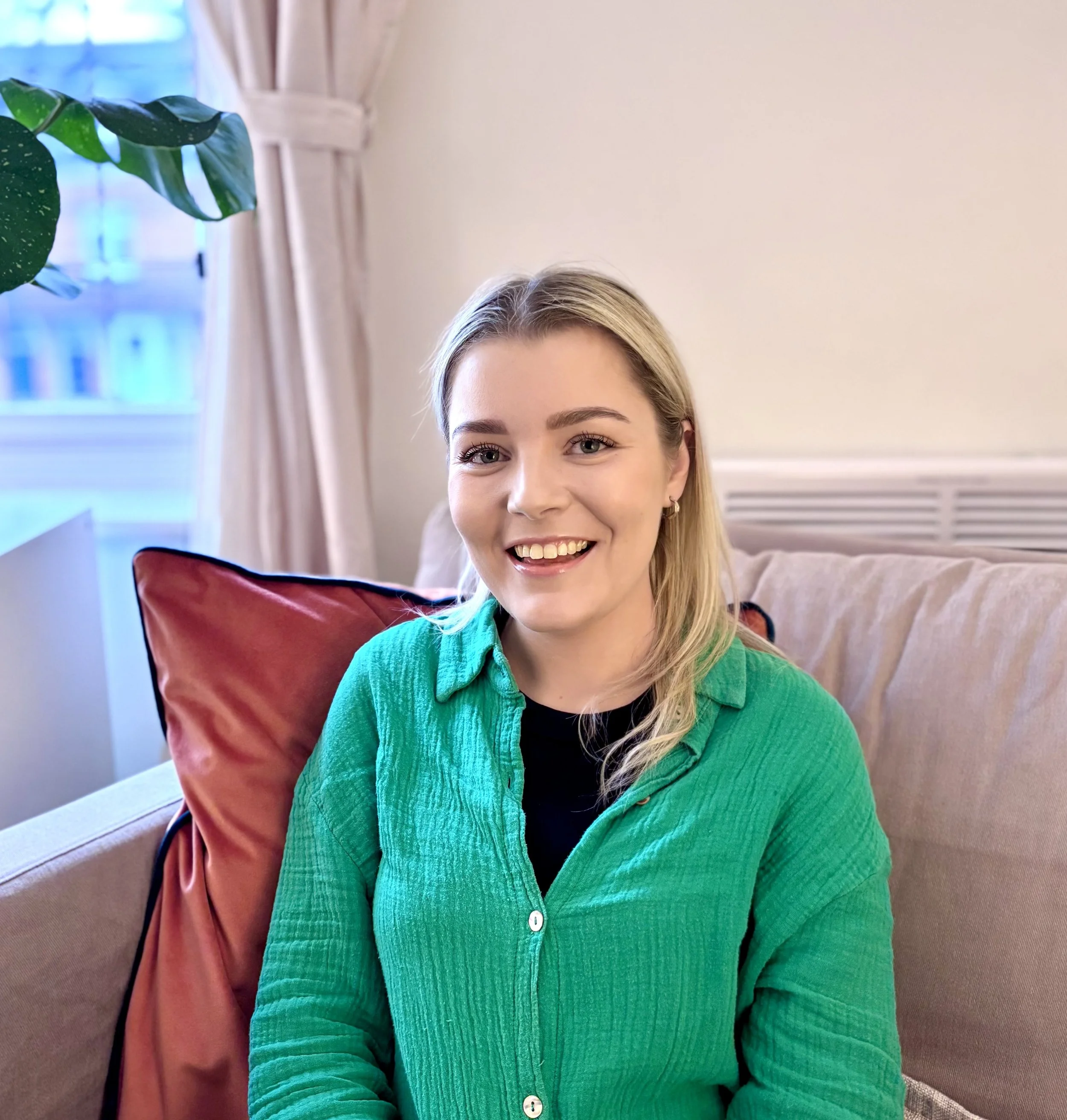 Team Member Shannon Rowe with blonde hair, smiling, wearing a green shirt, sitting on a beige sofa with a red cushion in a cozy living room.