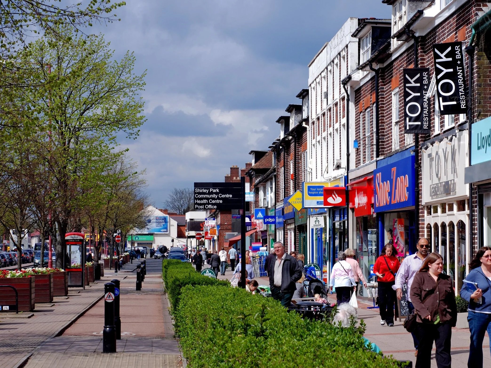 People walking along Shirley High Street with various store signs and a line of trees on the left. 