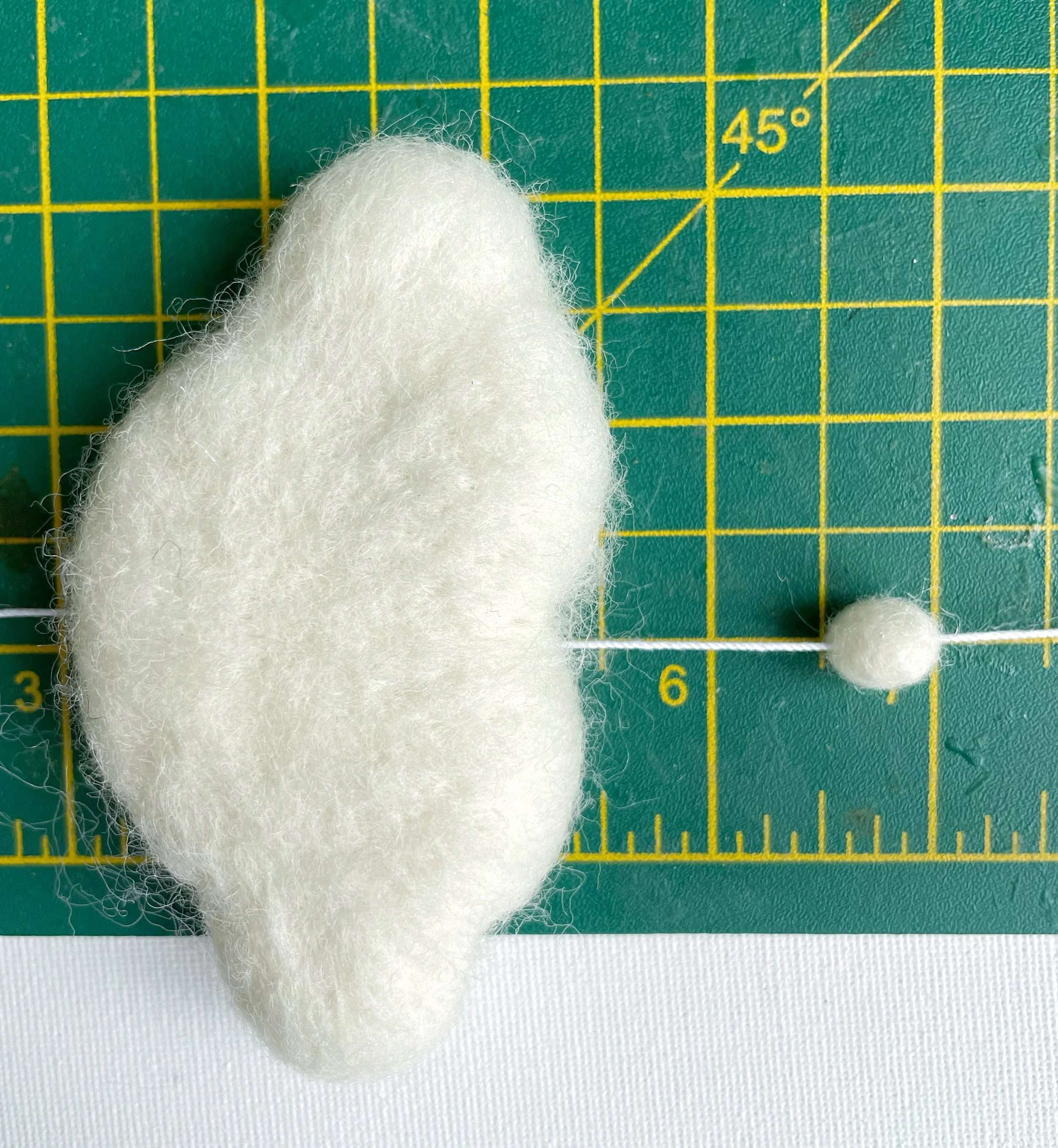 White wool felt cloud and white felt ball threaded onto white string. Measured on green cutting mat.