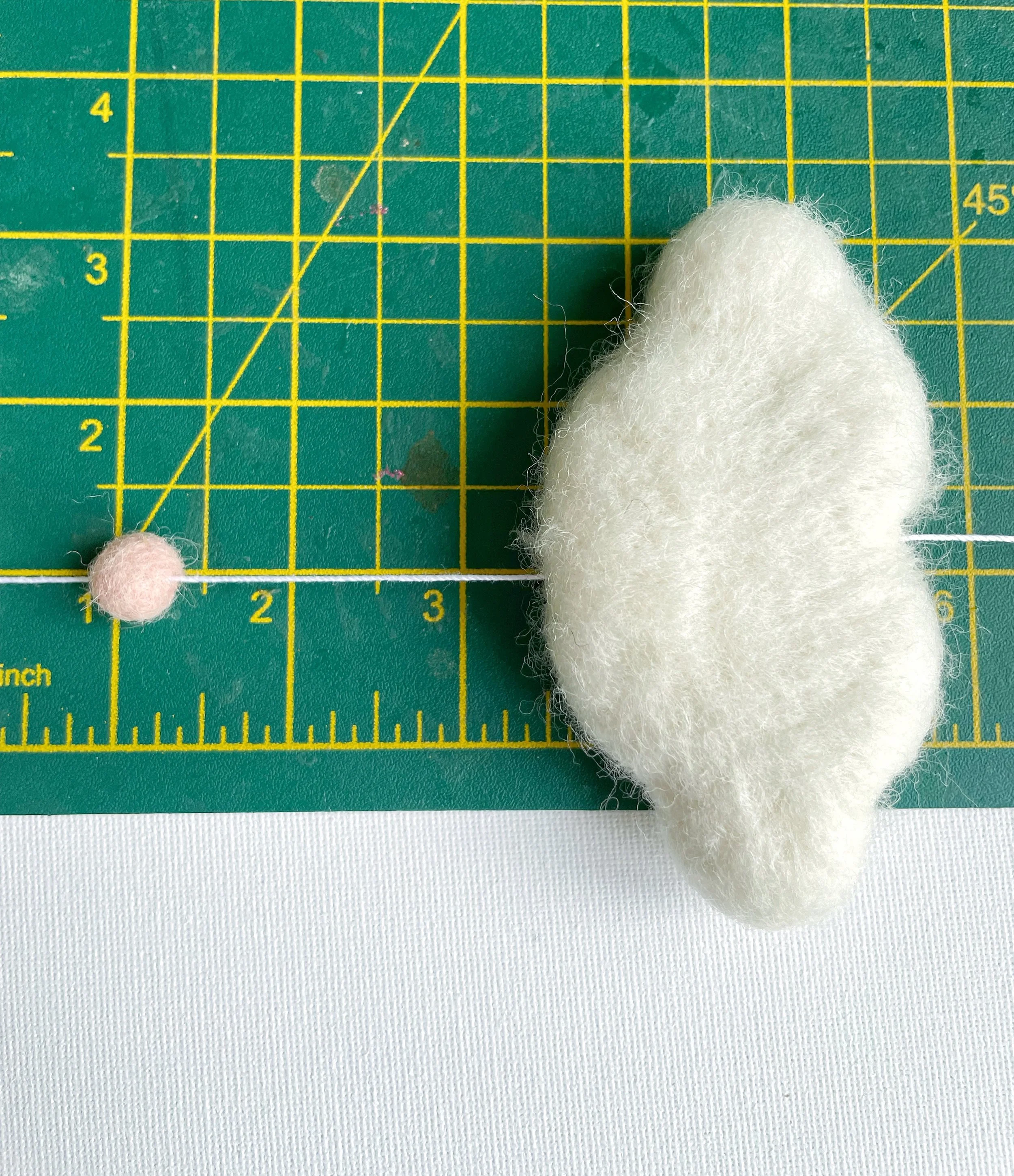 Pink felt ball and white wool felt cloud threaded onto white string. Measured on green cutting mat.