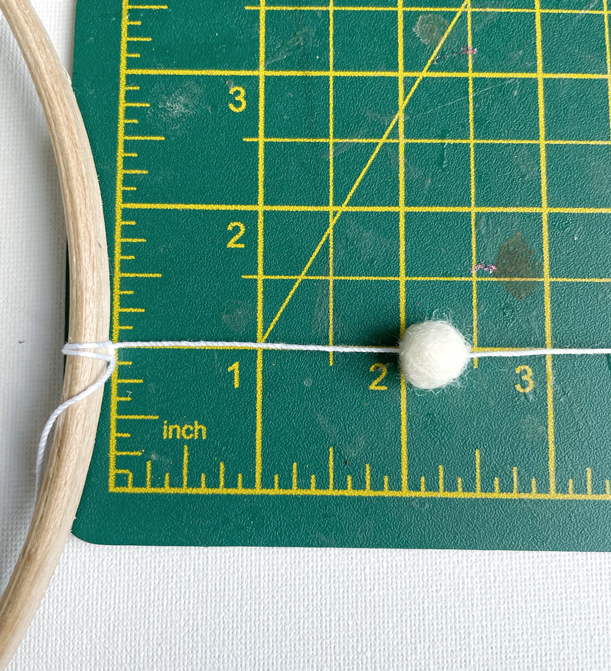 White felt ball threaded onto white string, shown at two inch point on green cutting mat. The sting is attached to a wooden hoop.