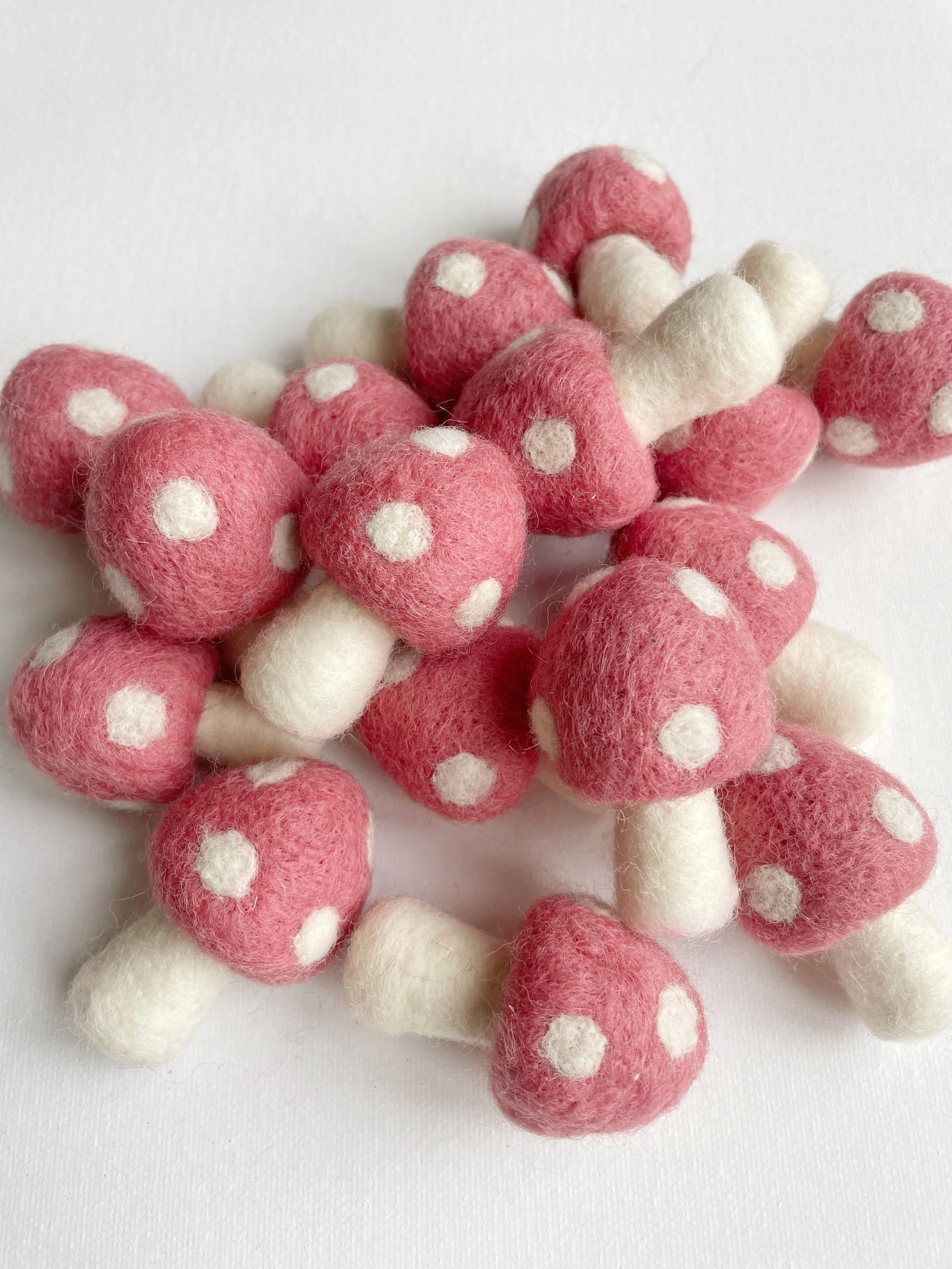 A collection of small felted pink mushrooms with white stems and white spots on their caps, scattered on a white background.