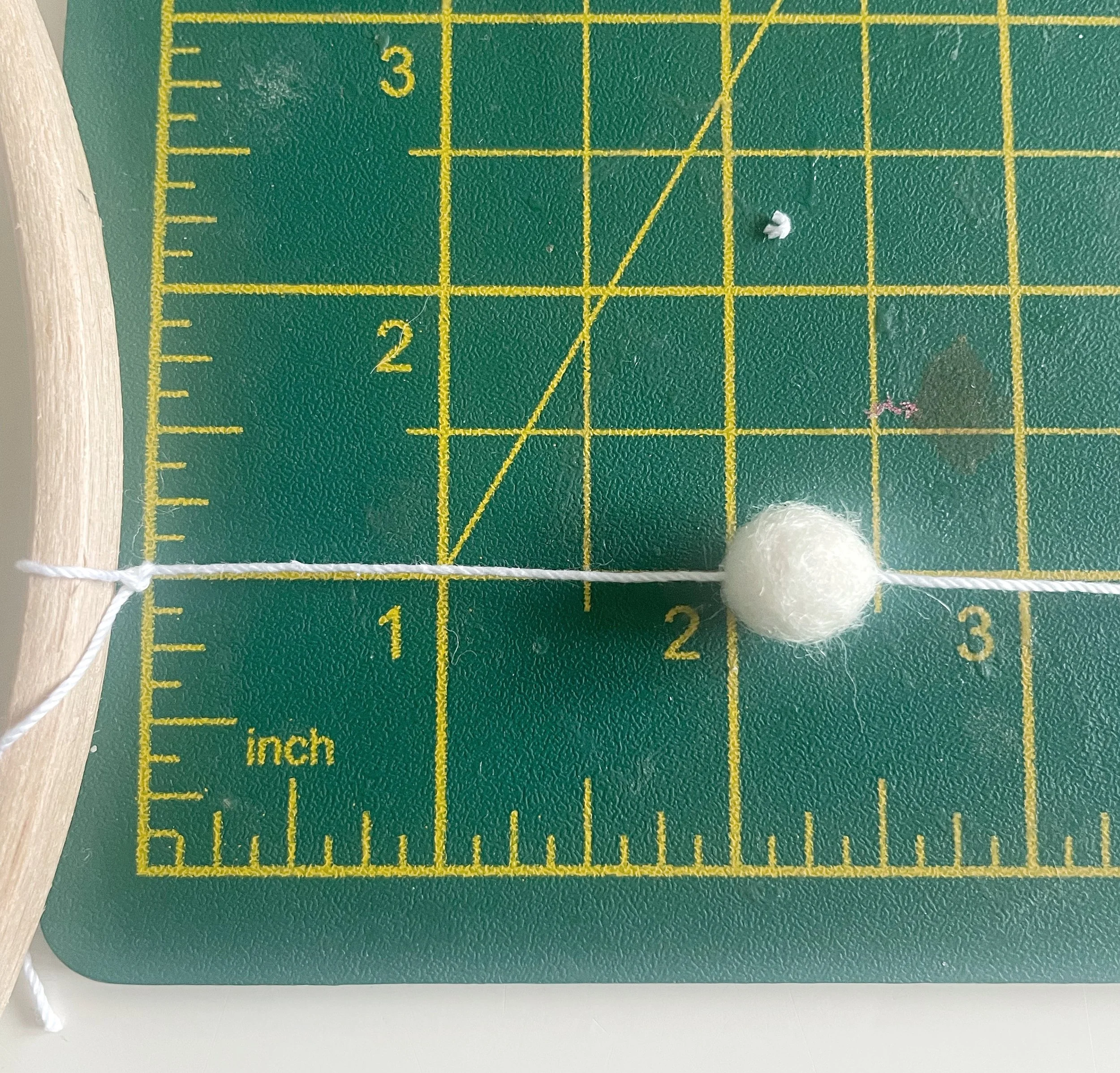 Green cutting mat with white felt ball threaded onto white string tied to wooden hoop. Felt ball is placed at 2 inch mark on mat.