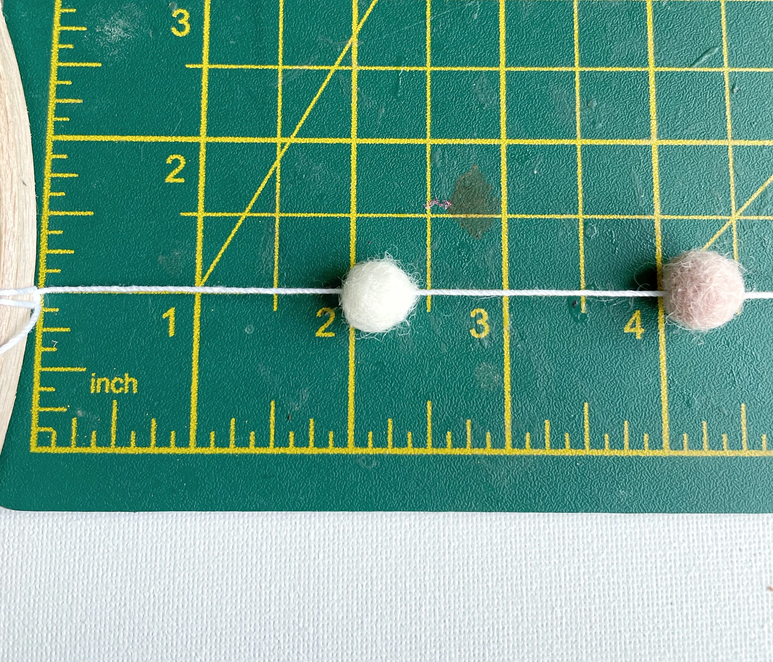 A pink and white felt ball threaded on to a white piece of string. Placed two inches apart, measured on a green cutting mat.